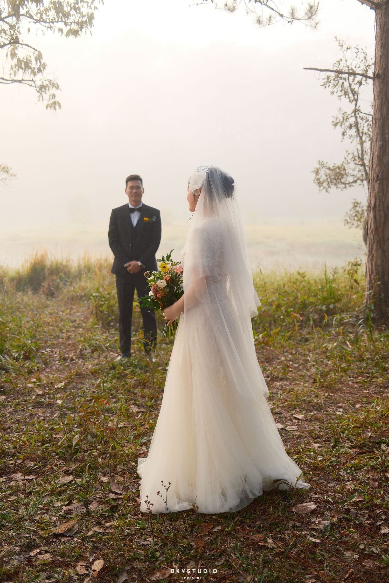 Bride in white gown holds bouquet facing groom in misty woodland landscape during outdoor wedding shoot.