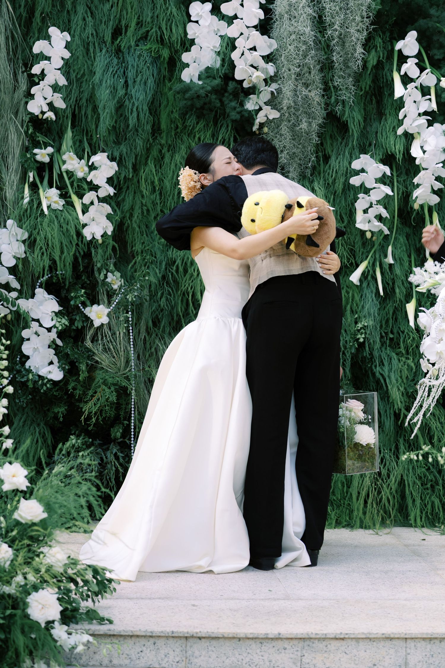 Bride and groom share an emotional hug at the altar surrounded by white orchids and greenery.