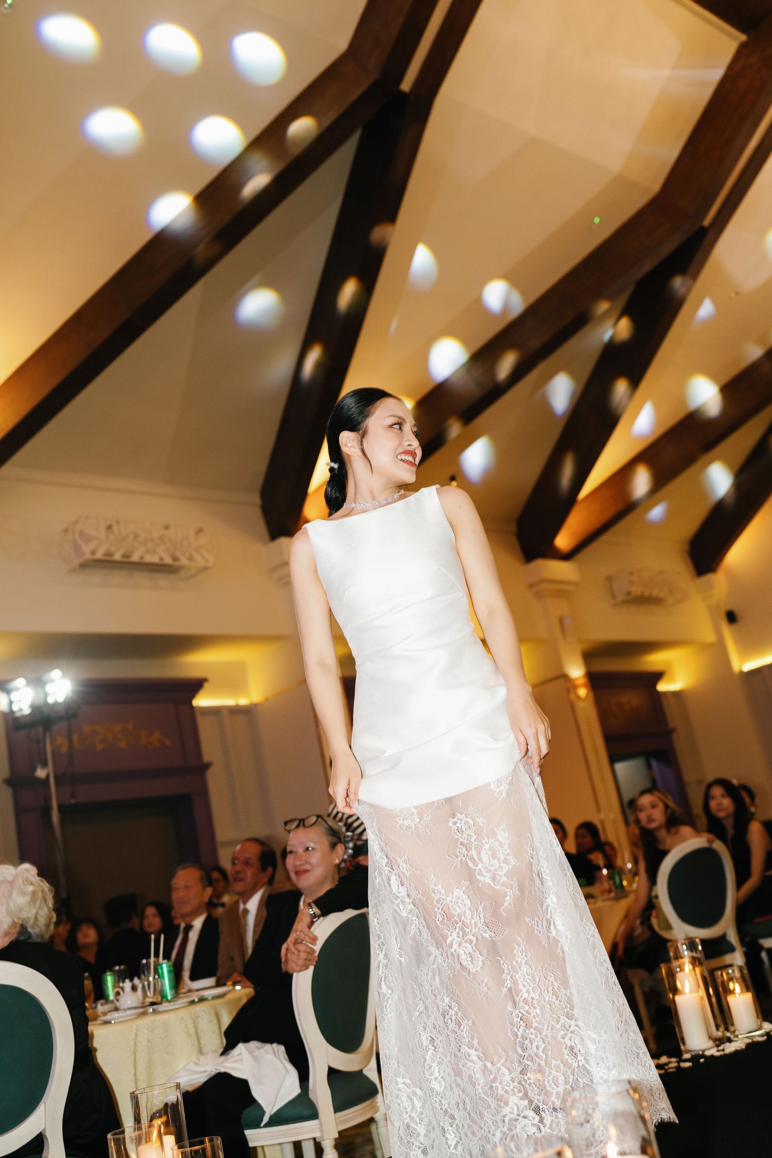 Bride in a sleeveless white gown with lace skirt walks elegantly through a warmly lit ballroom during her wedding reception.