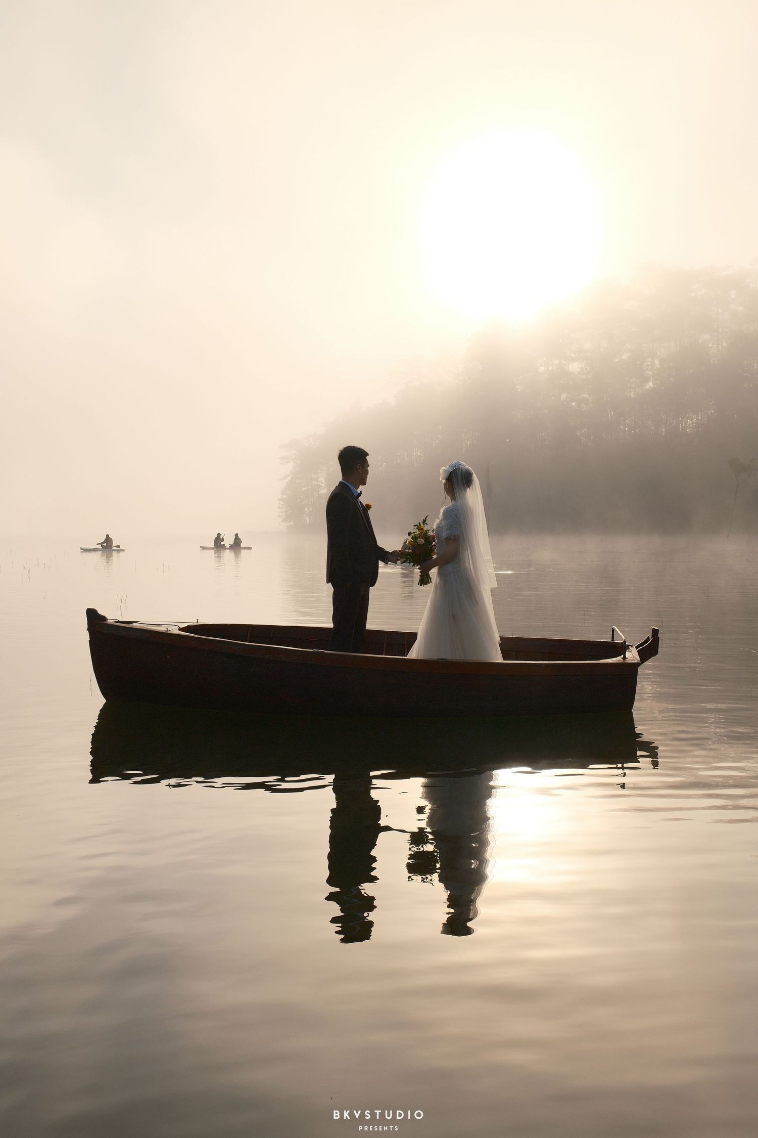 Bride and groom stand in wooden boat on misty lake at sunrise, silhouetted against golden light.