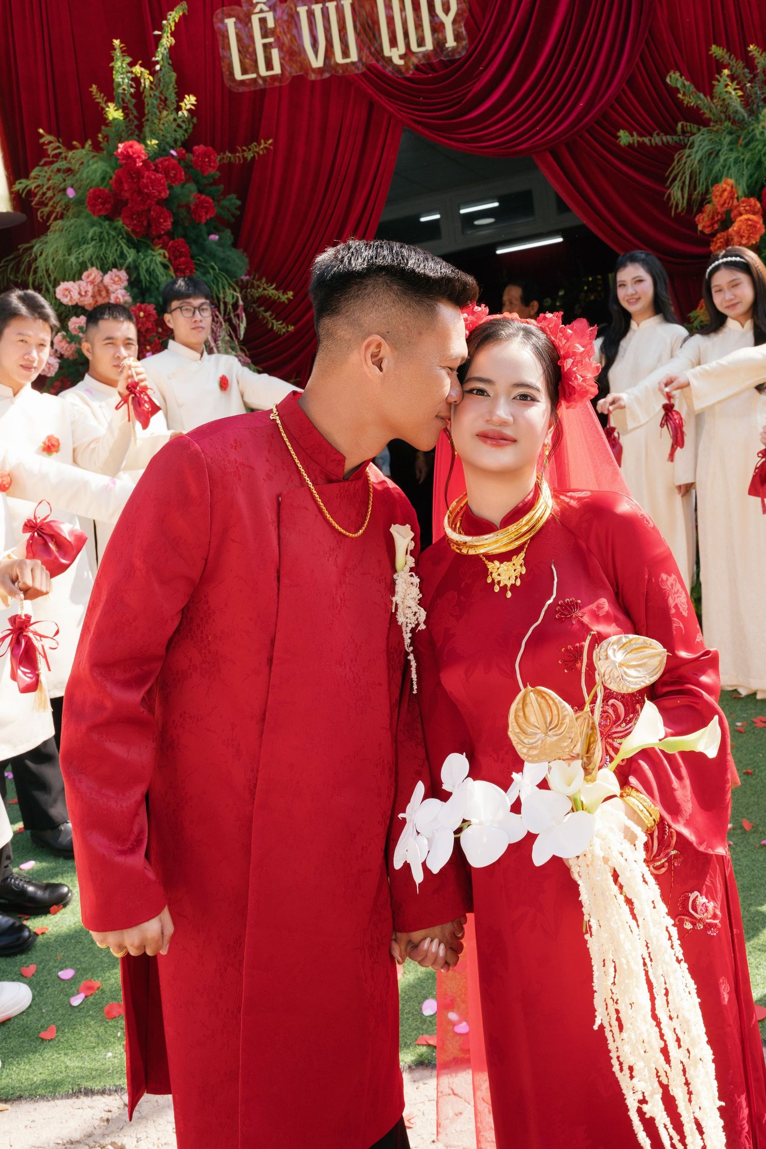 Vietnamese couple in red ao dai traditional wedding attire at a Le Vu Quy ceremony, groom kissing bride's cheek.