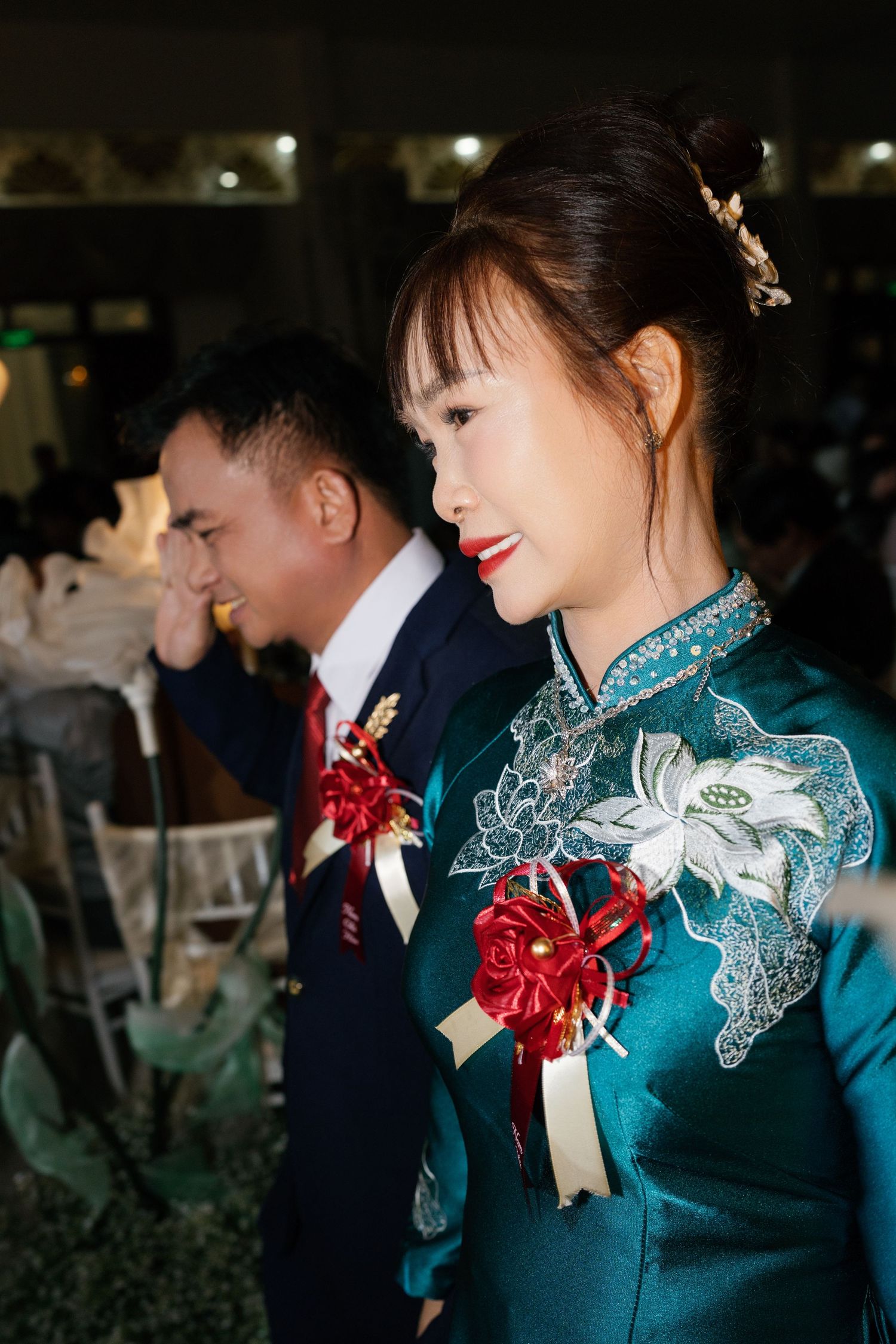 Vietnamese bride in teal ao dai with red floral pin smiles during traditional wedding banquet ceremony.