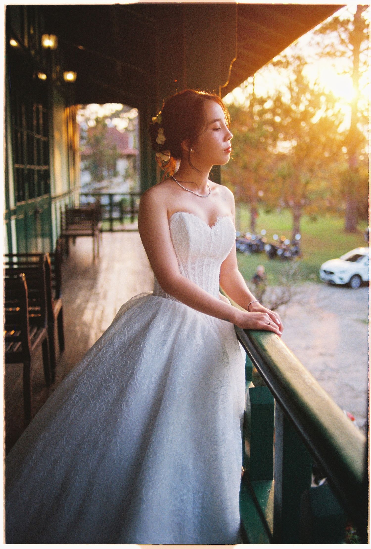 A bride in a strapless white ball gown stands on a balcony at golden hour, gazing into the distance.