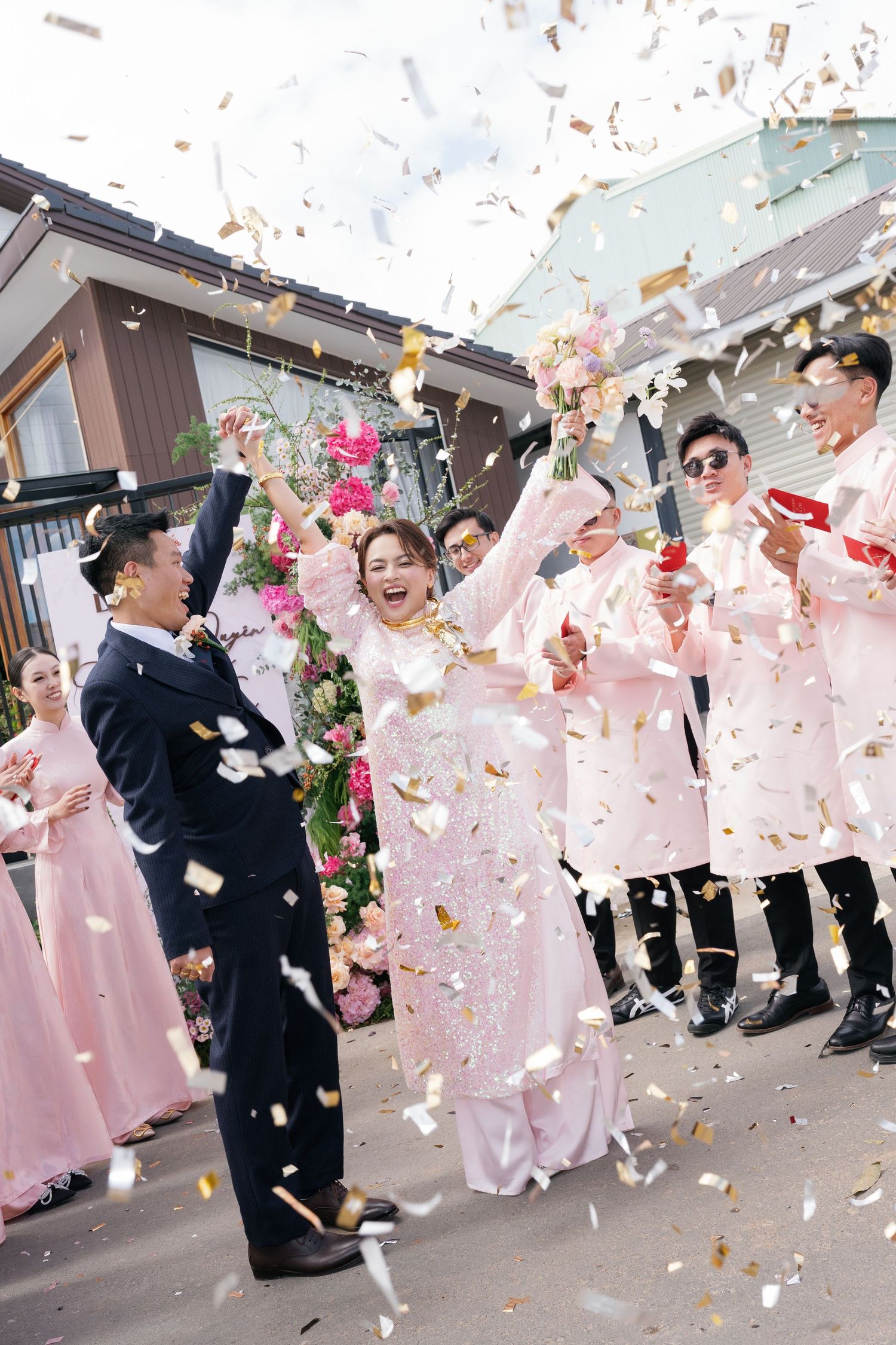 A joyful bride and groom celebrate with confetti falling as wedding party members cheer outdoors.