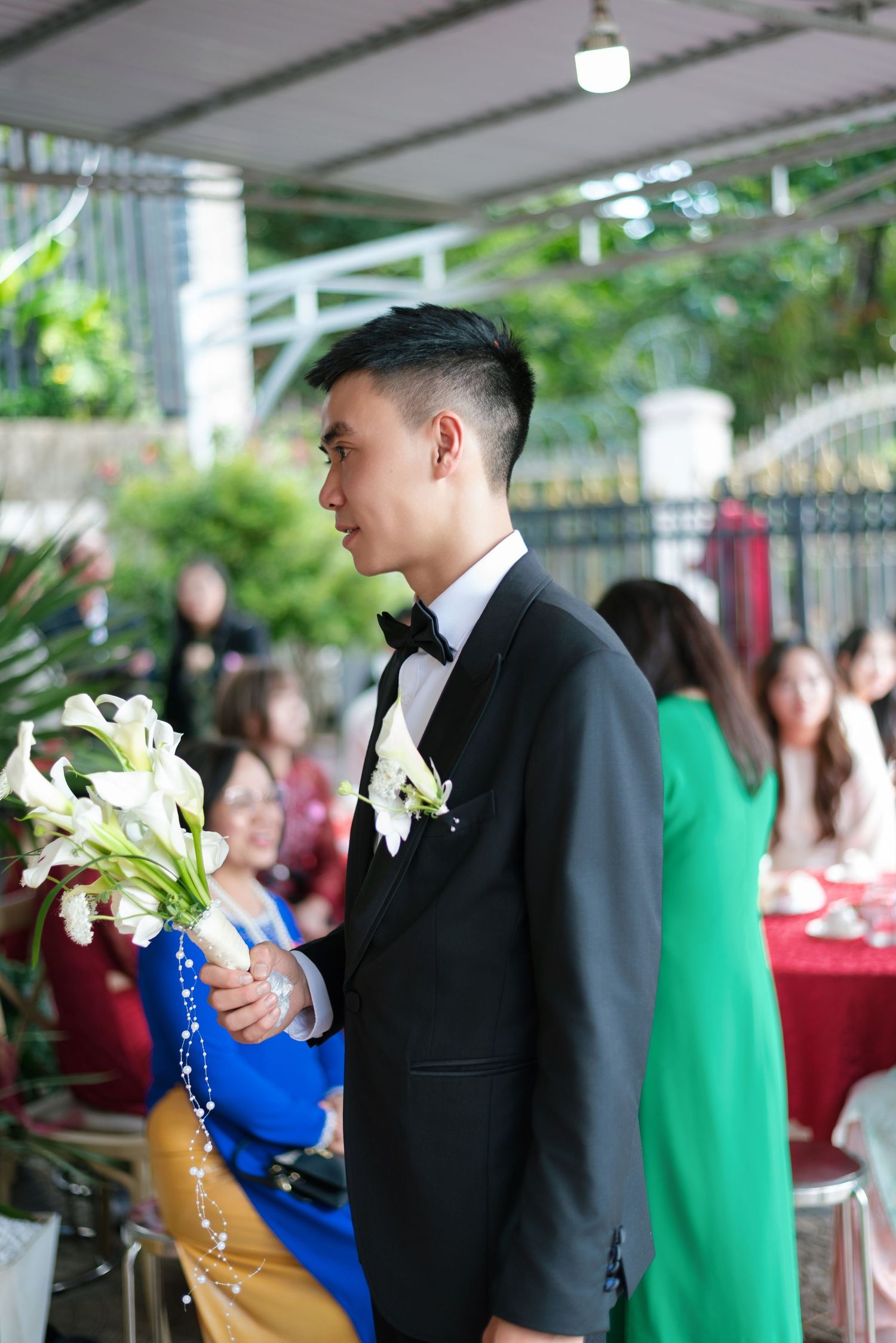 Groom in black tuxedo holding white calla lily bouquet at outdoor wedding ceremony with guests seated nearby.
