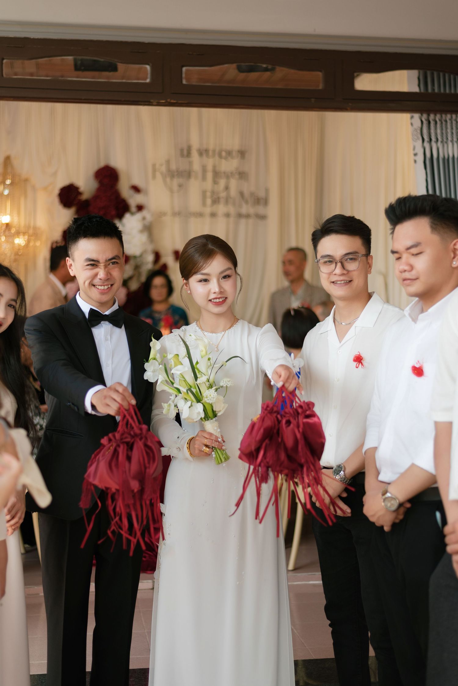 Vietnamese wedding ceremony with bride in white ao dai holding flowers, groom in tuxedo, and guests holding red decorations.