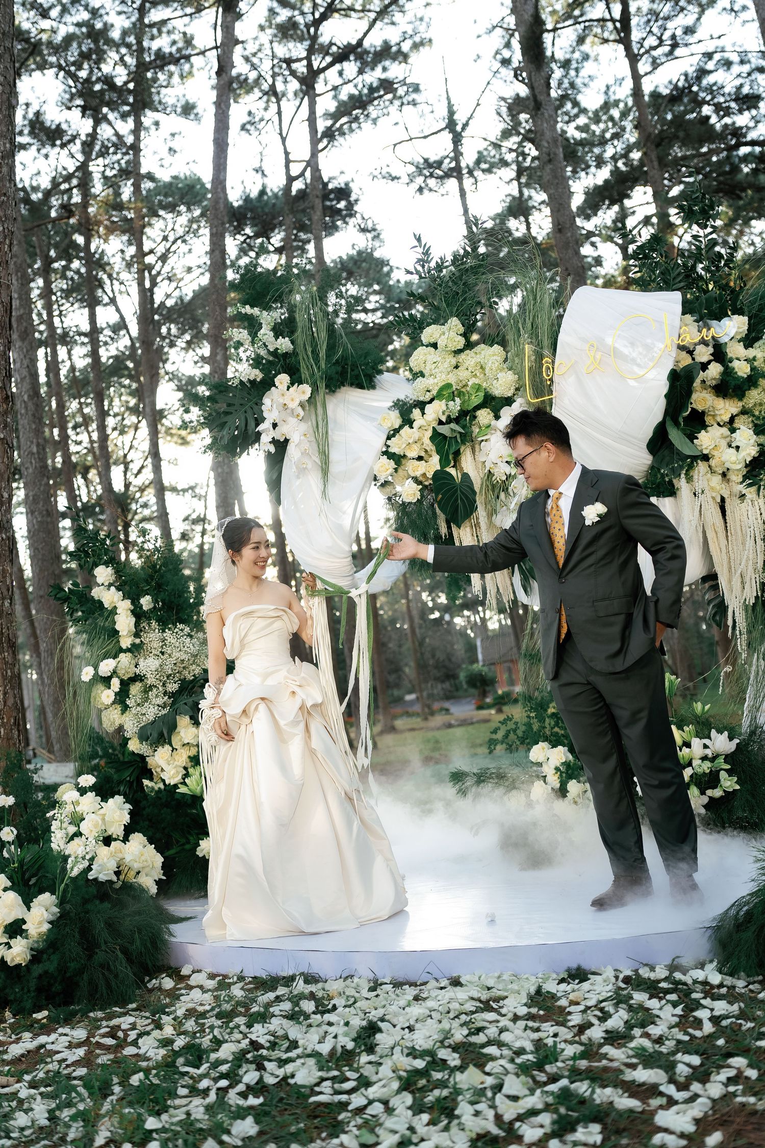 Bride in strapless gown and groom in dark suit celebrate at a floral outdoor wedding altar surrounded by pine trees.
