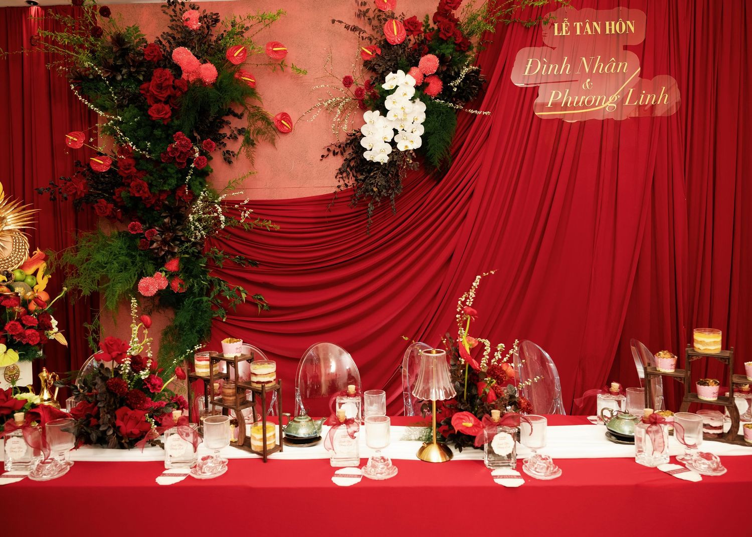 Red wedding reception table with floral arrangements, candles, and a backdrop reading Dinh Nhan & Phuong Linh.