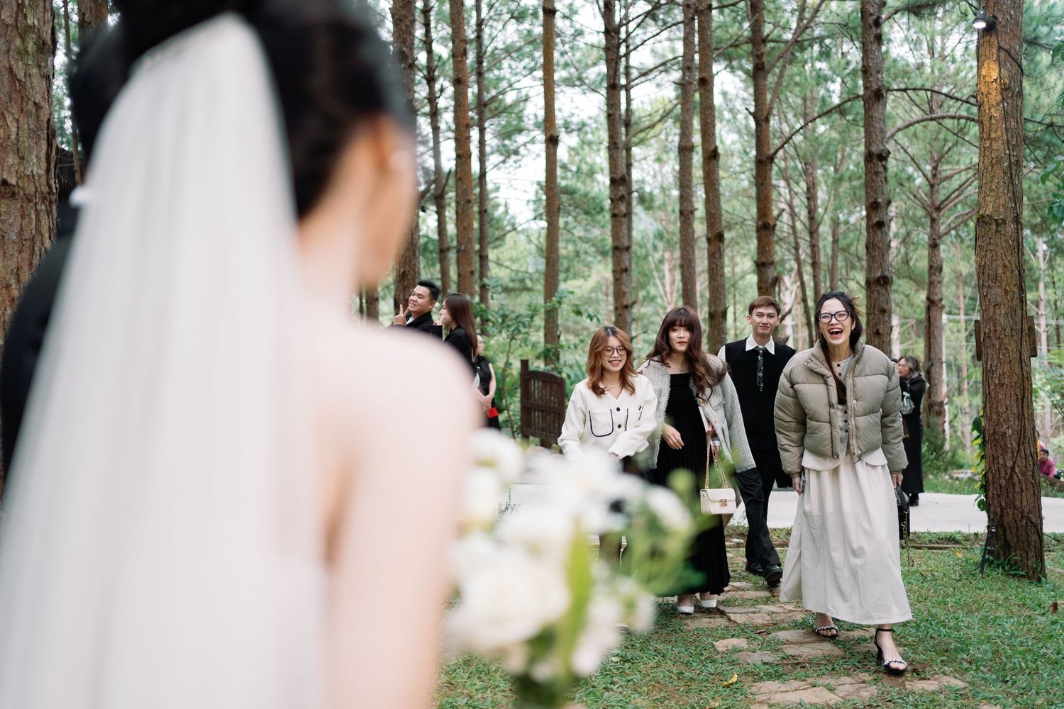 Outdoor forest wedding ceremony with bride holding bouquet as guests walk through pine trees in elegant attire.