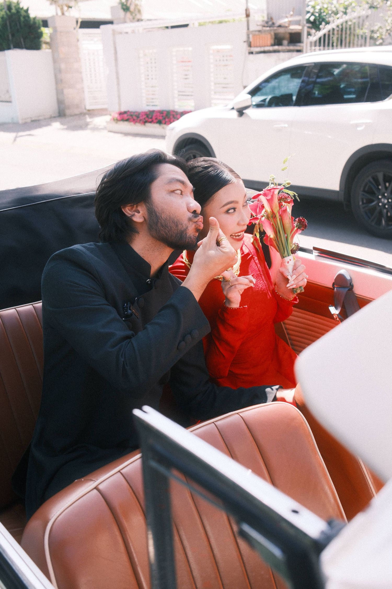 Couple in vintage convertible car, man in black kissing woman in red holding pink flowers on sunny day.