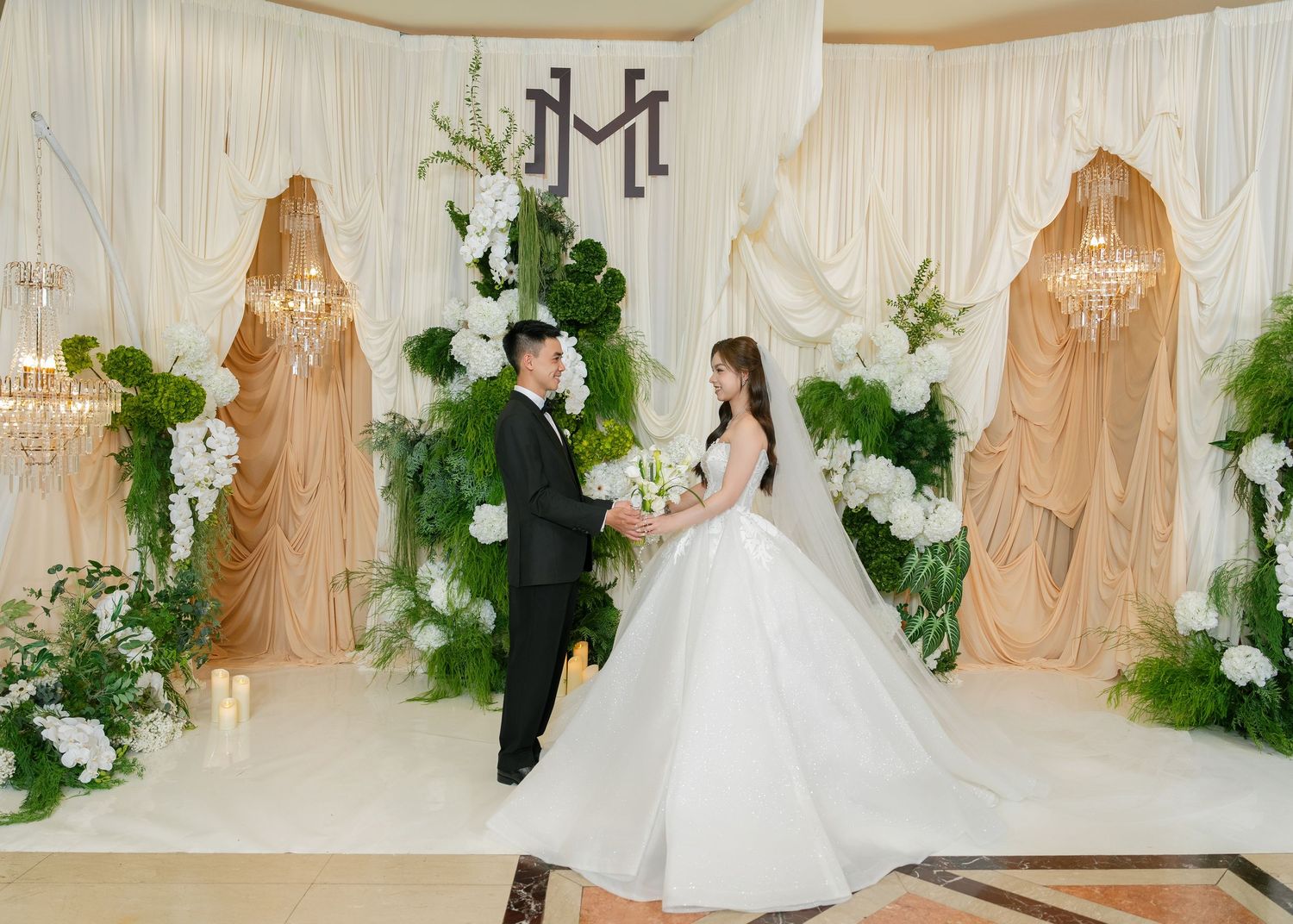 A bride in a white ball gown and groom in a black suit stand at an elegant floral wedding altar with greenery.