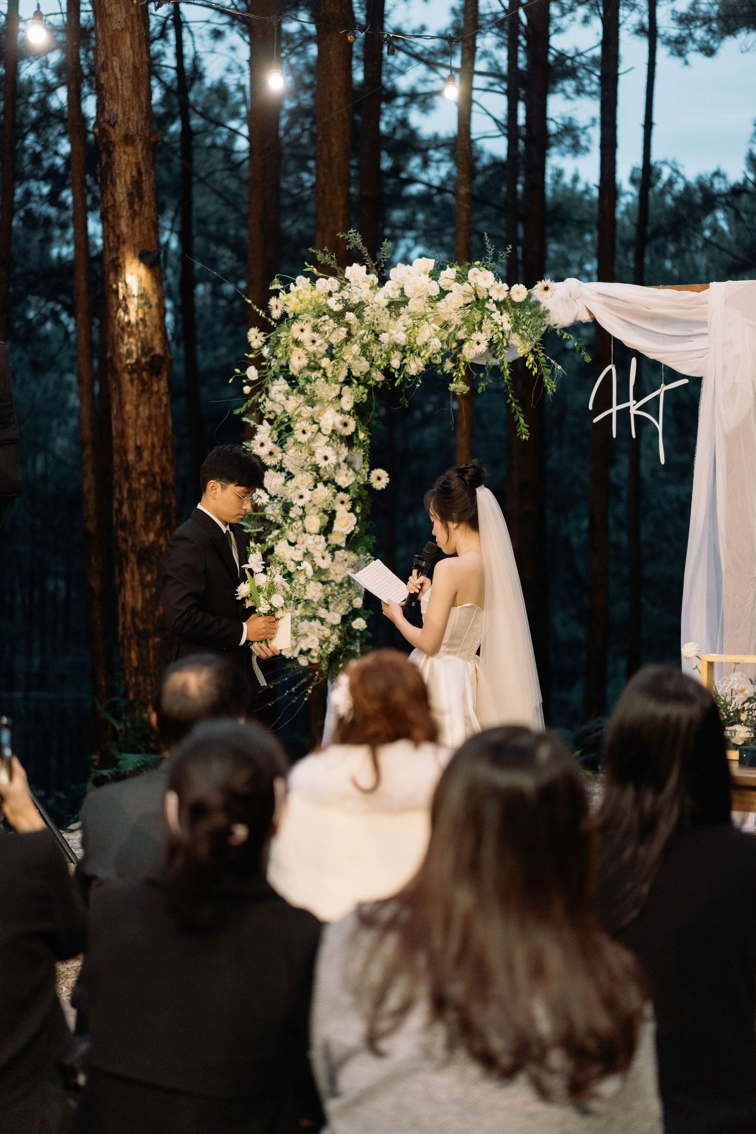 Couple exchanging vows at an outdoor forest wedding ceremony with a white floral arch and seated guests.