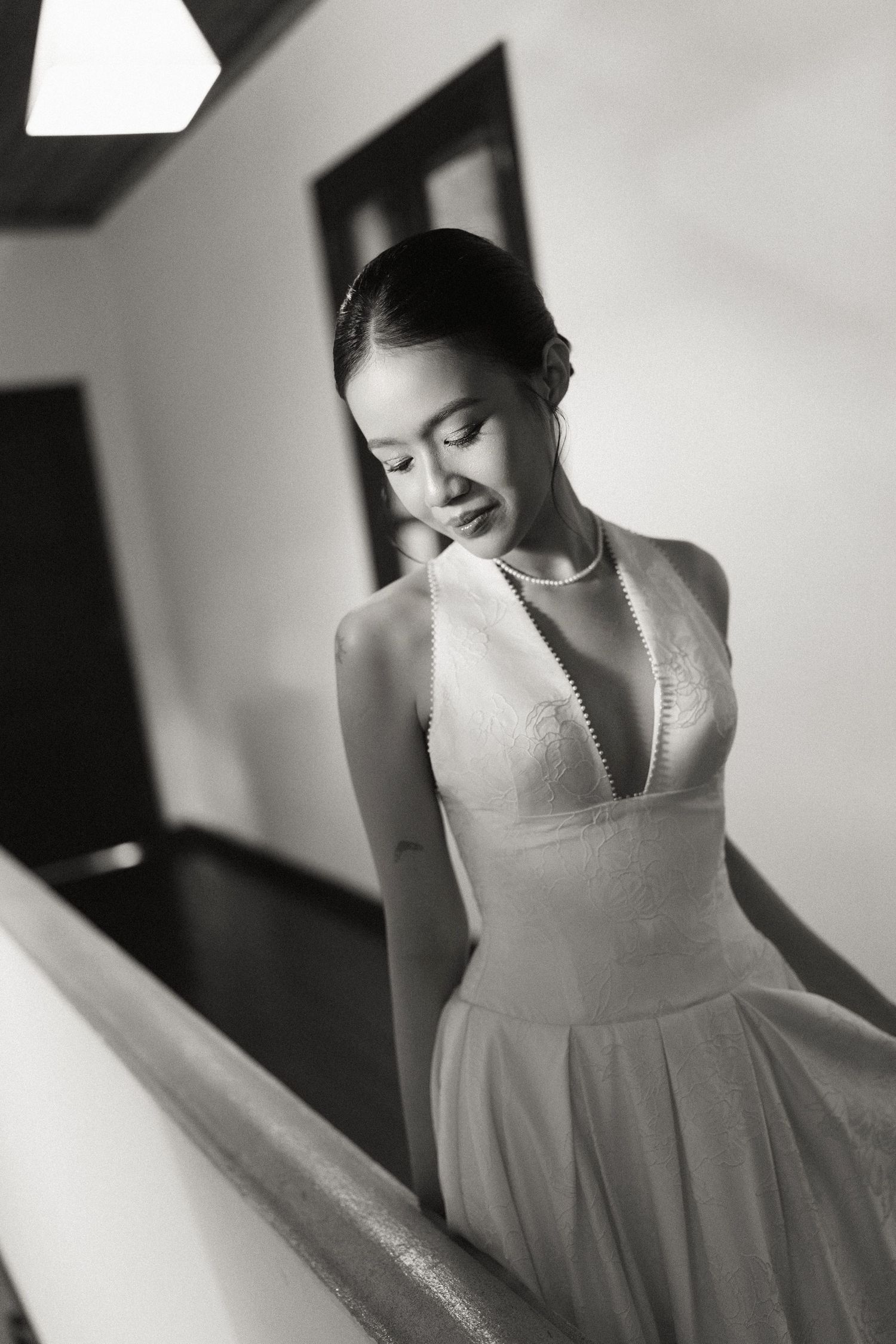 Black and white portrait of elegant bride in halter wedding gown with pearl necklace on staircase.