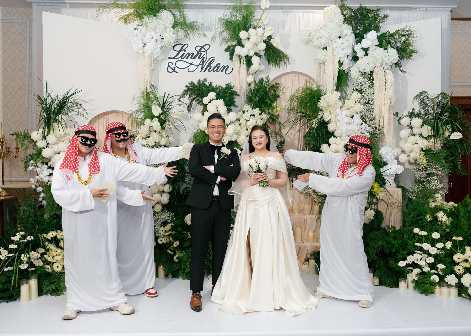 A bride and groom pose with guests dressed in traditional Arab attire at an elegant floral wedding reception backdrop.