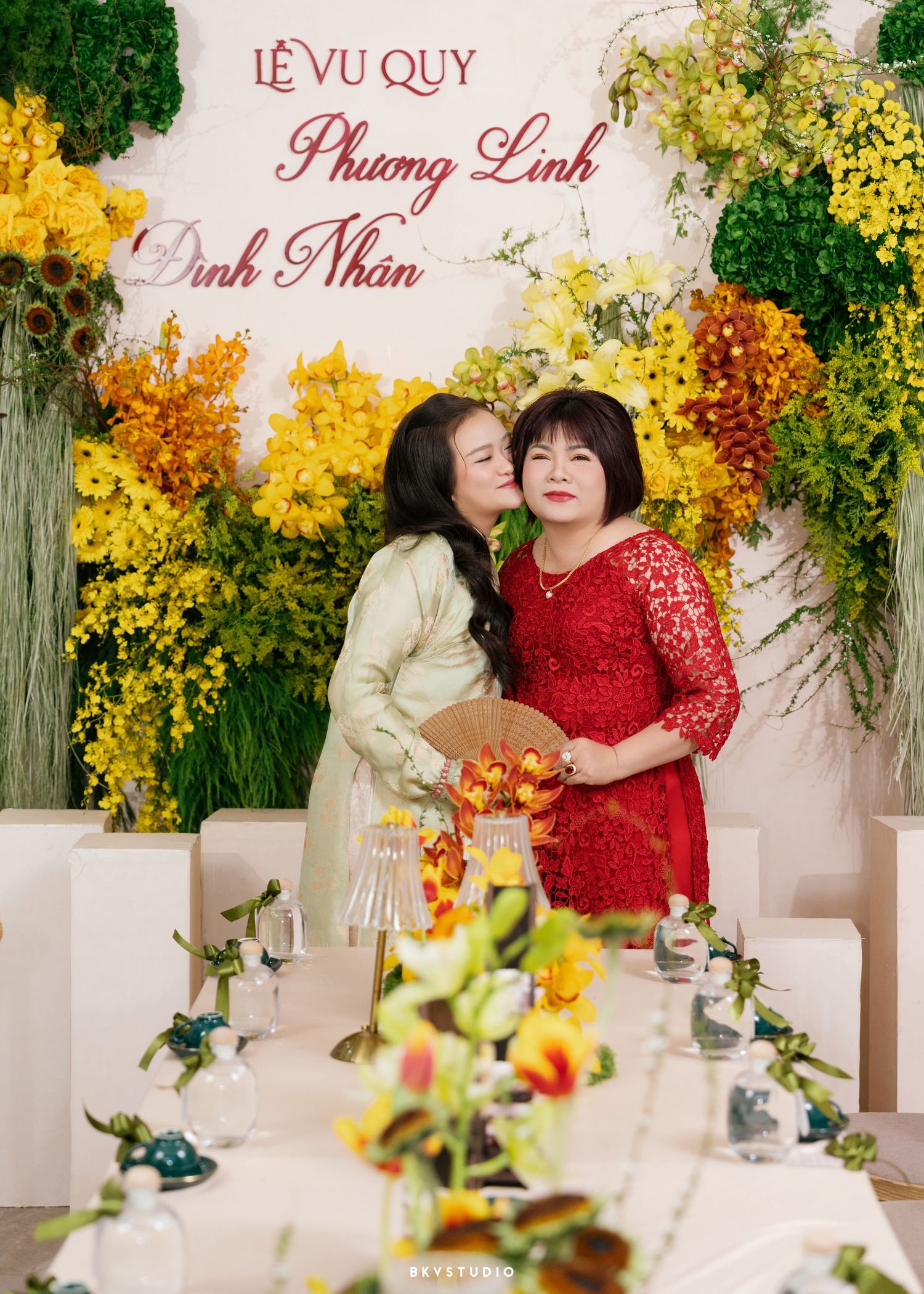 Two women share a tender moment at a Vietnamese wedding ceremony, surrounded by vibrant yellow floral decorations.