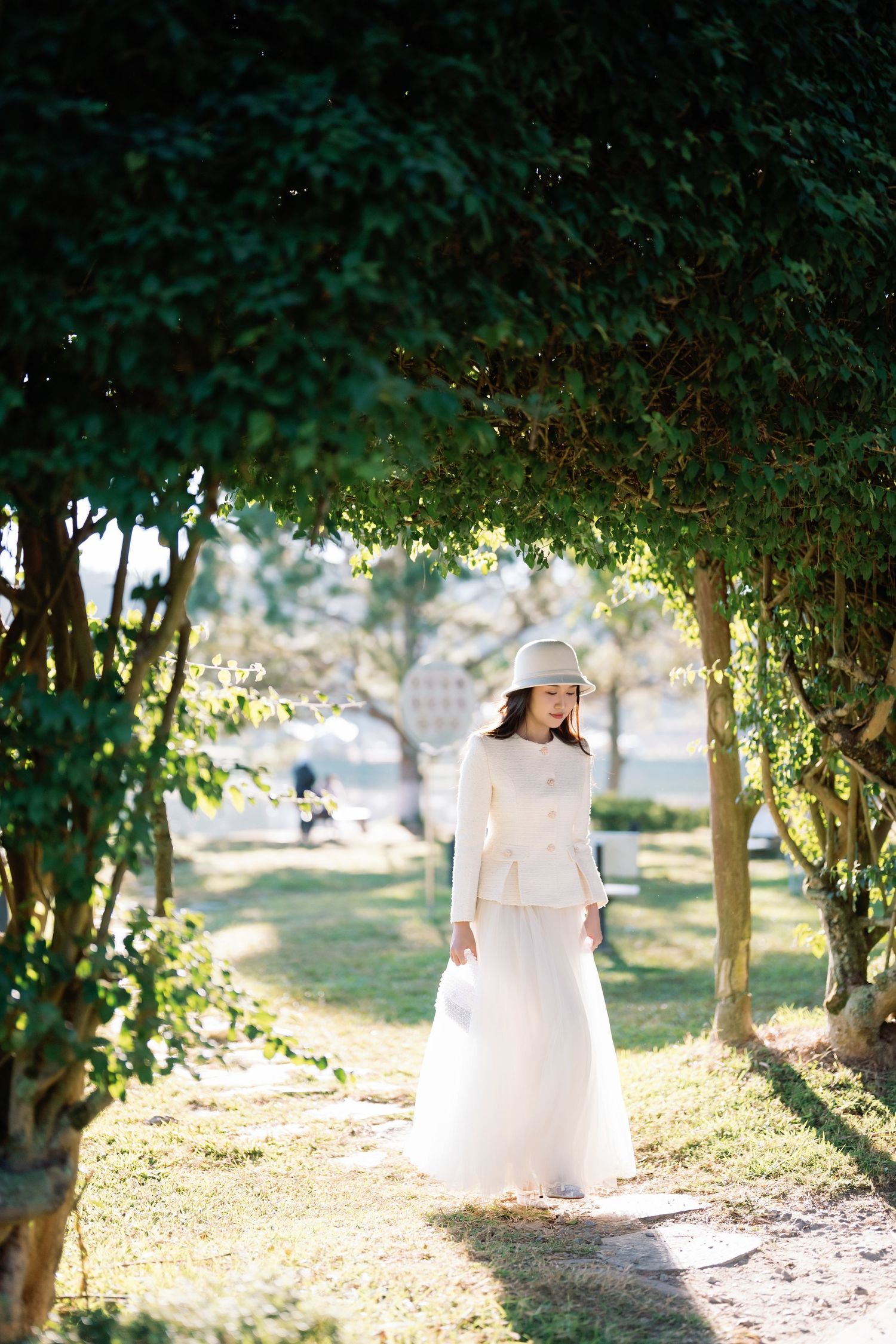 A woman in a white dress and hat stands elegantly under a lush green archway in a sunlit park.