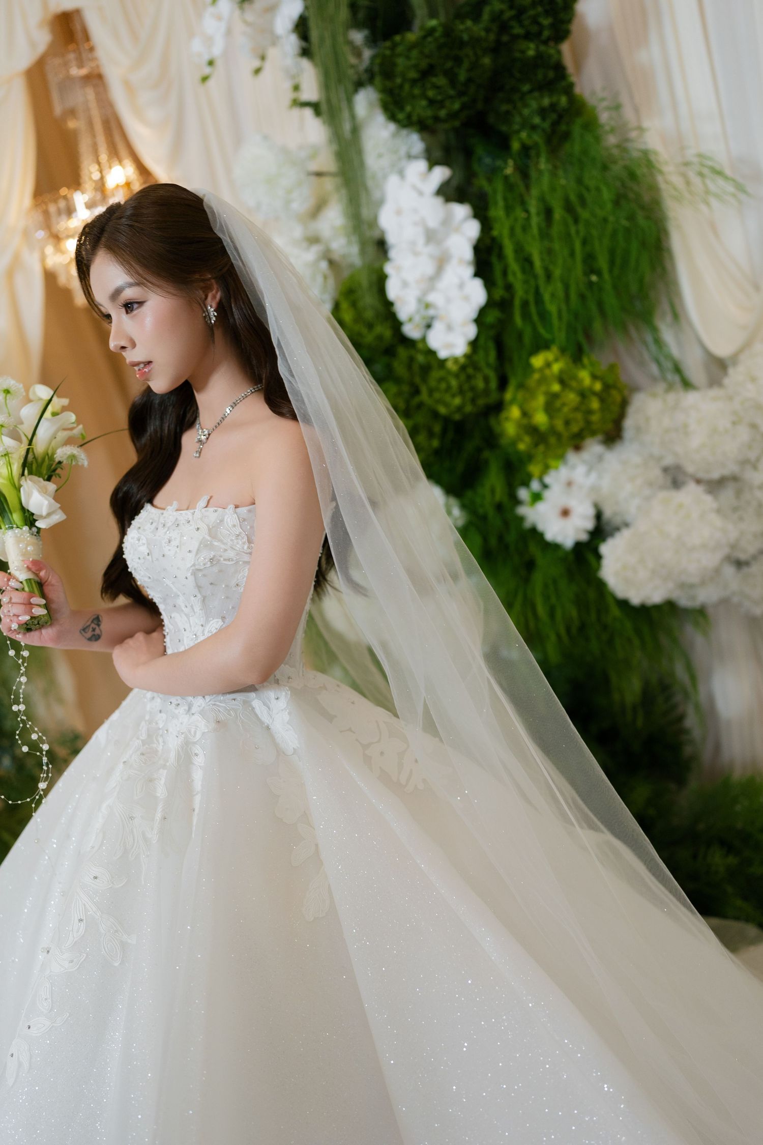Elegant bride in a strapless white ball gown with veil, holding bouquet near floral backdrop.
