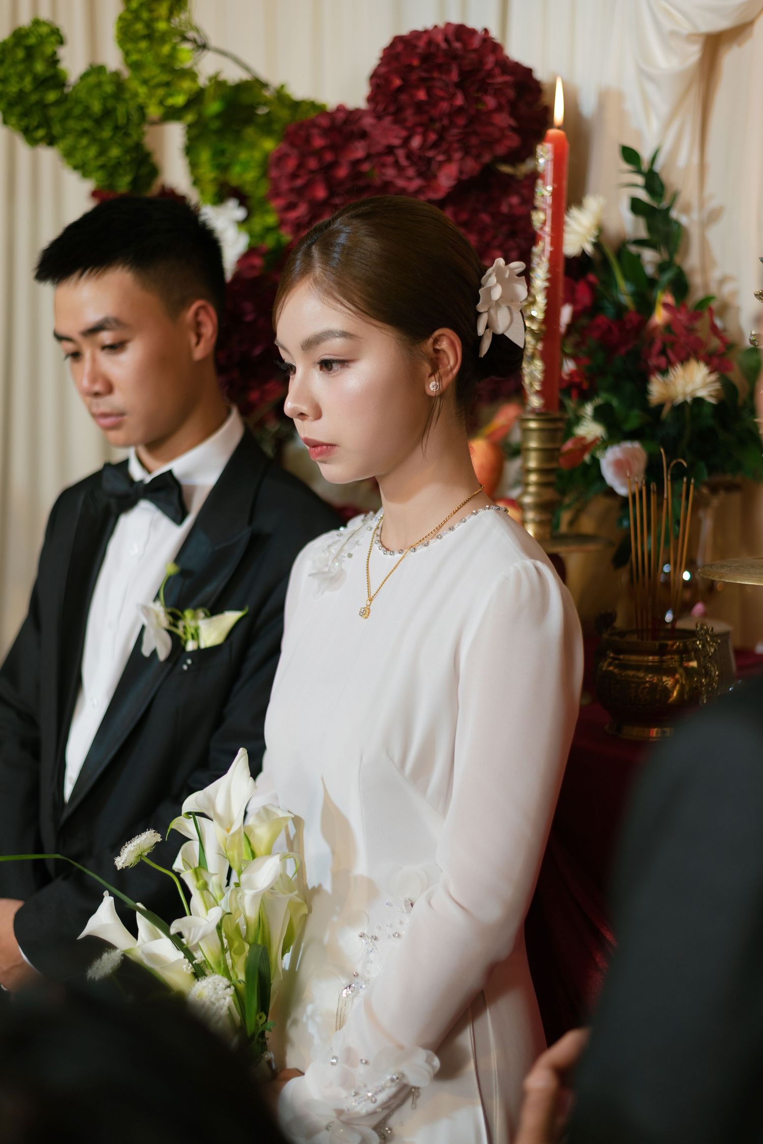 A bride in white dress holding lily bouquet stands beside groom in tuxedo during a traditional wedding ceremony.
