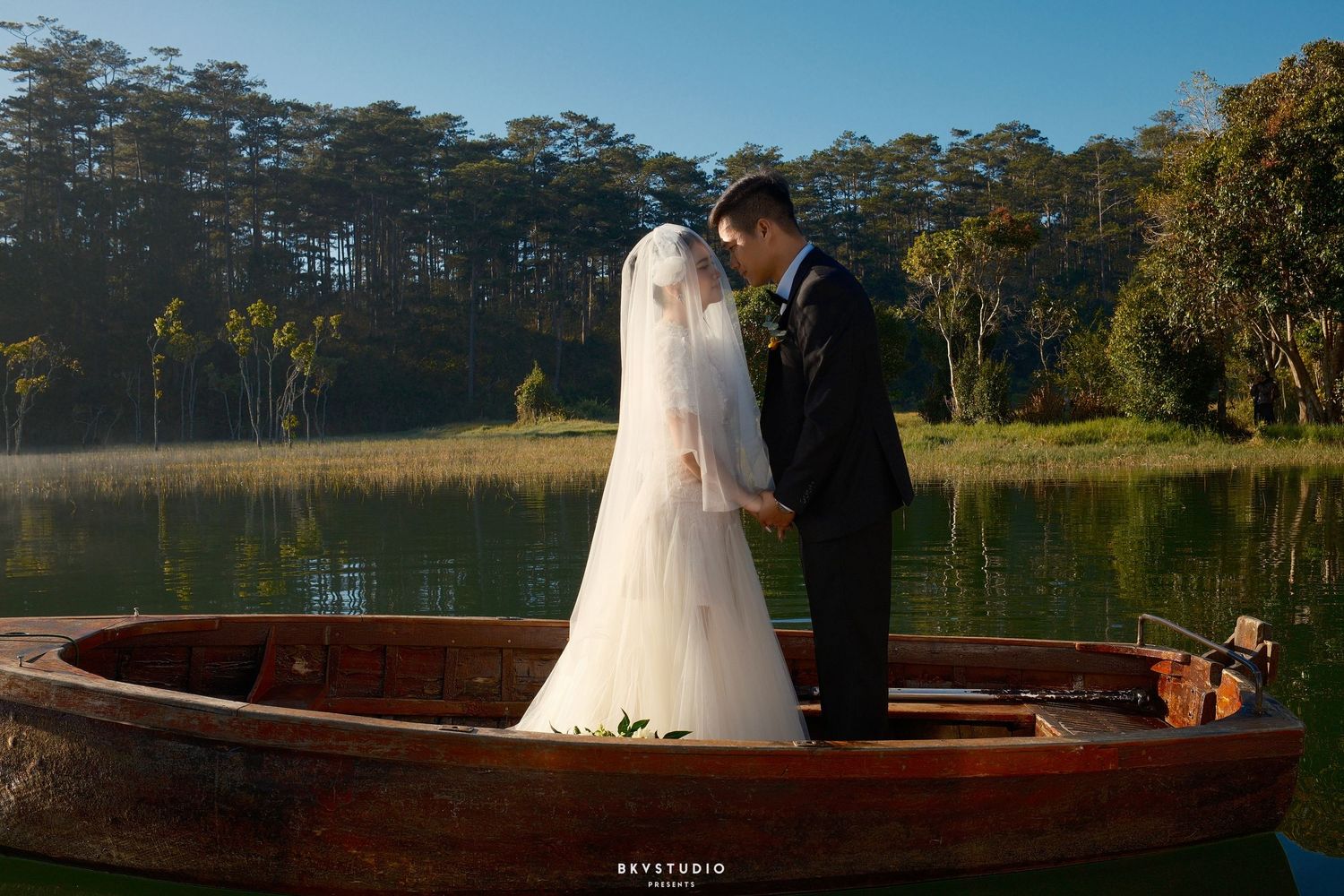 Bride and groom share a romantic moment standing in a wooden boat on a calm misty lake.