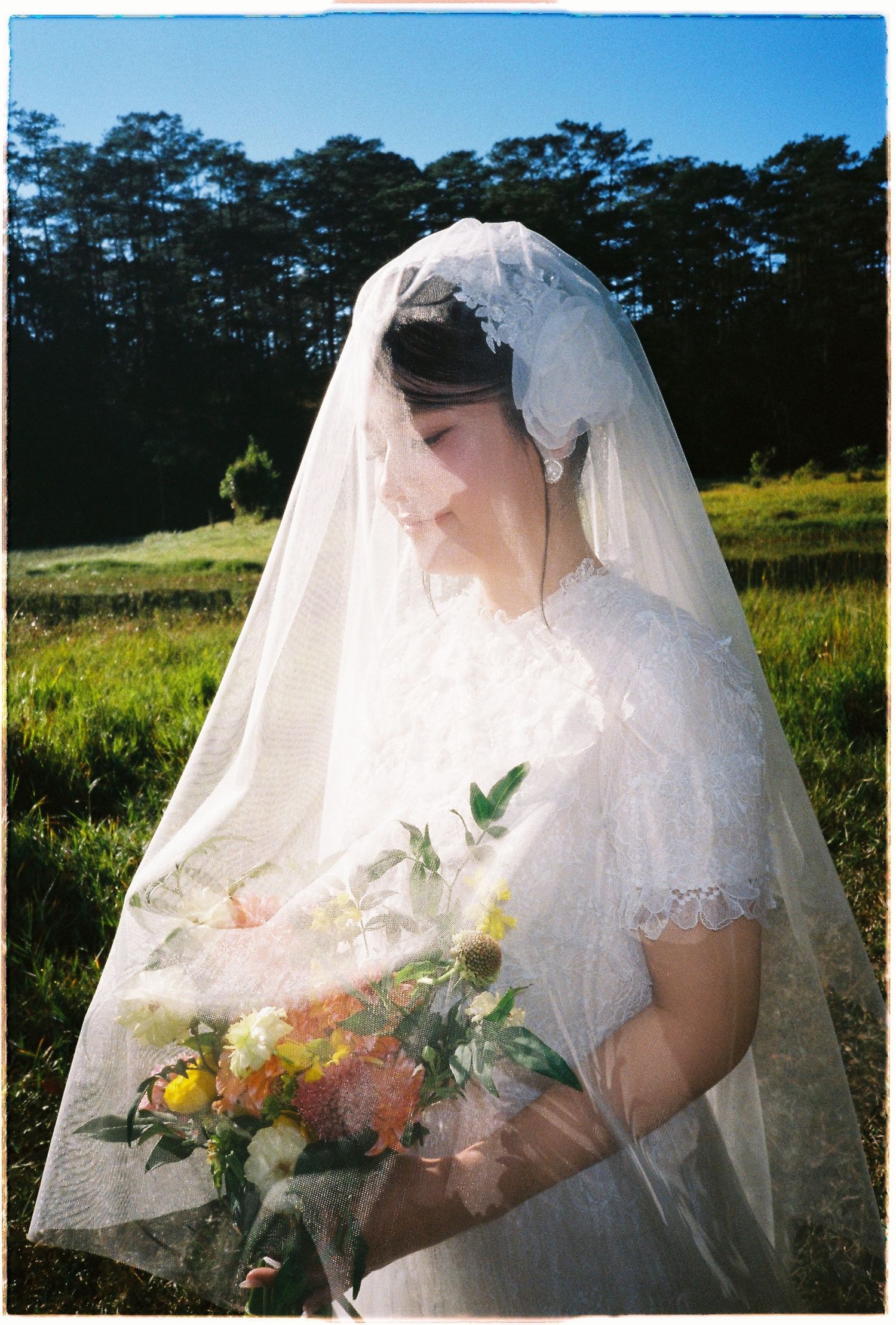 Bride in white lace dress and veil holds colorful bouquet while standing in sunny green meadow.