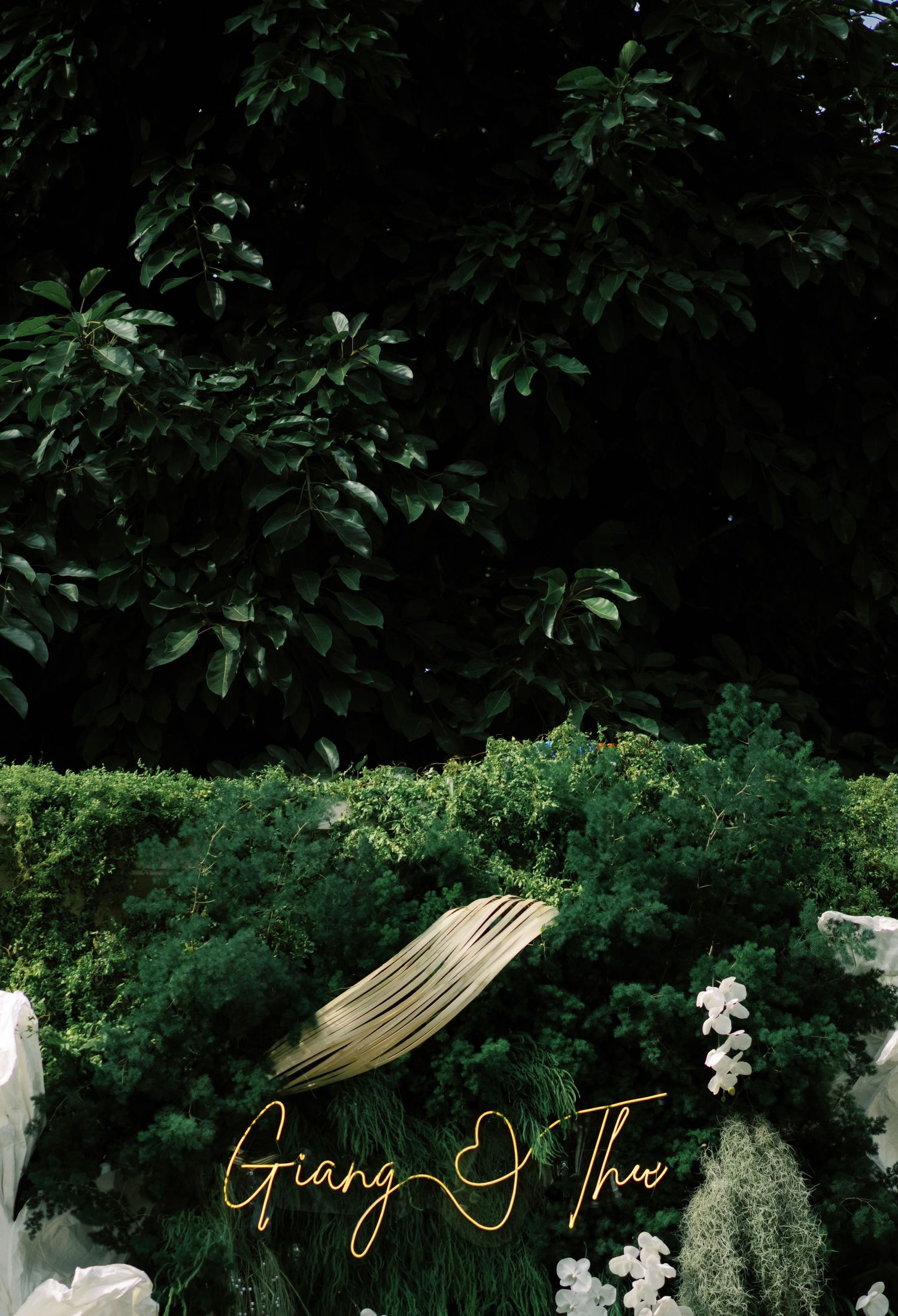 Lush green garden wedding backdrop with gold neon sign reading Giang and Thu surrounded by white flowers.