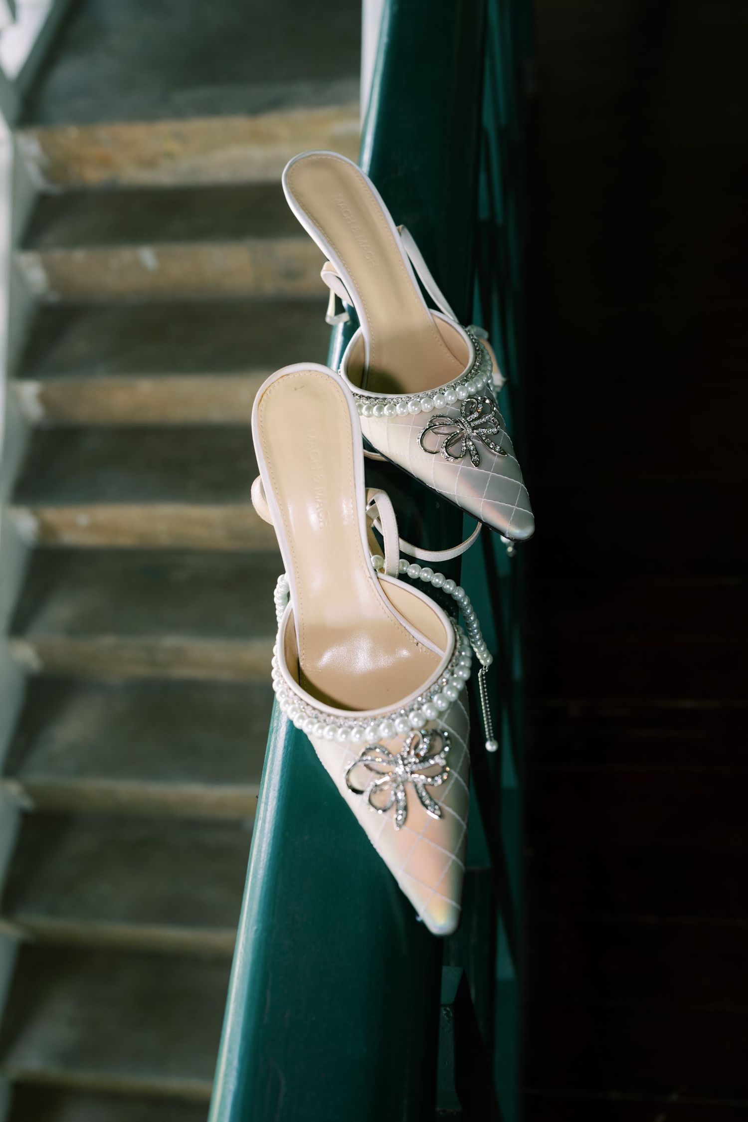 Elegant bridal heels with crystal embellishments and pearl details hanging on a dark green staircase railing.
