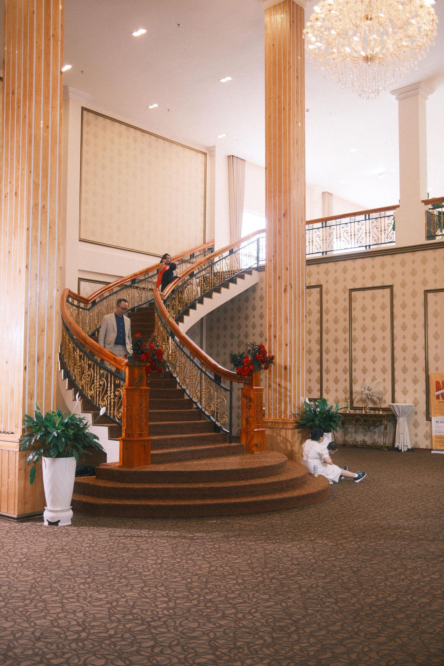 Elegant hotel lobby featuring a grand curved staircase with ornate railings, tall columns, chandelier, and decorative holiday greenery.