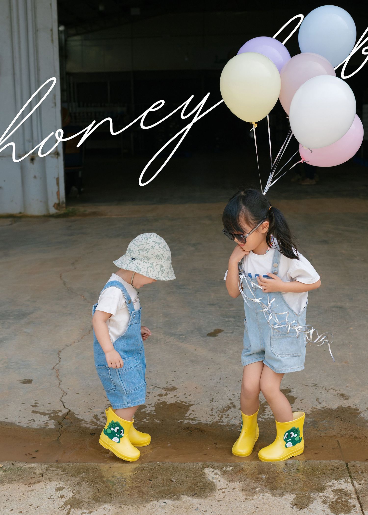 Two young children in denim overalls and yellow rain boots hold pastel balloons outdoors.