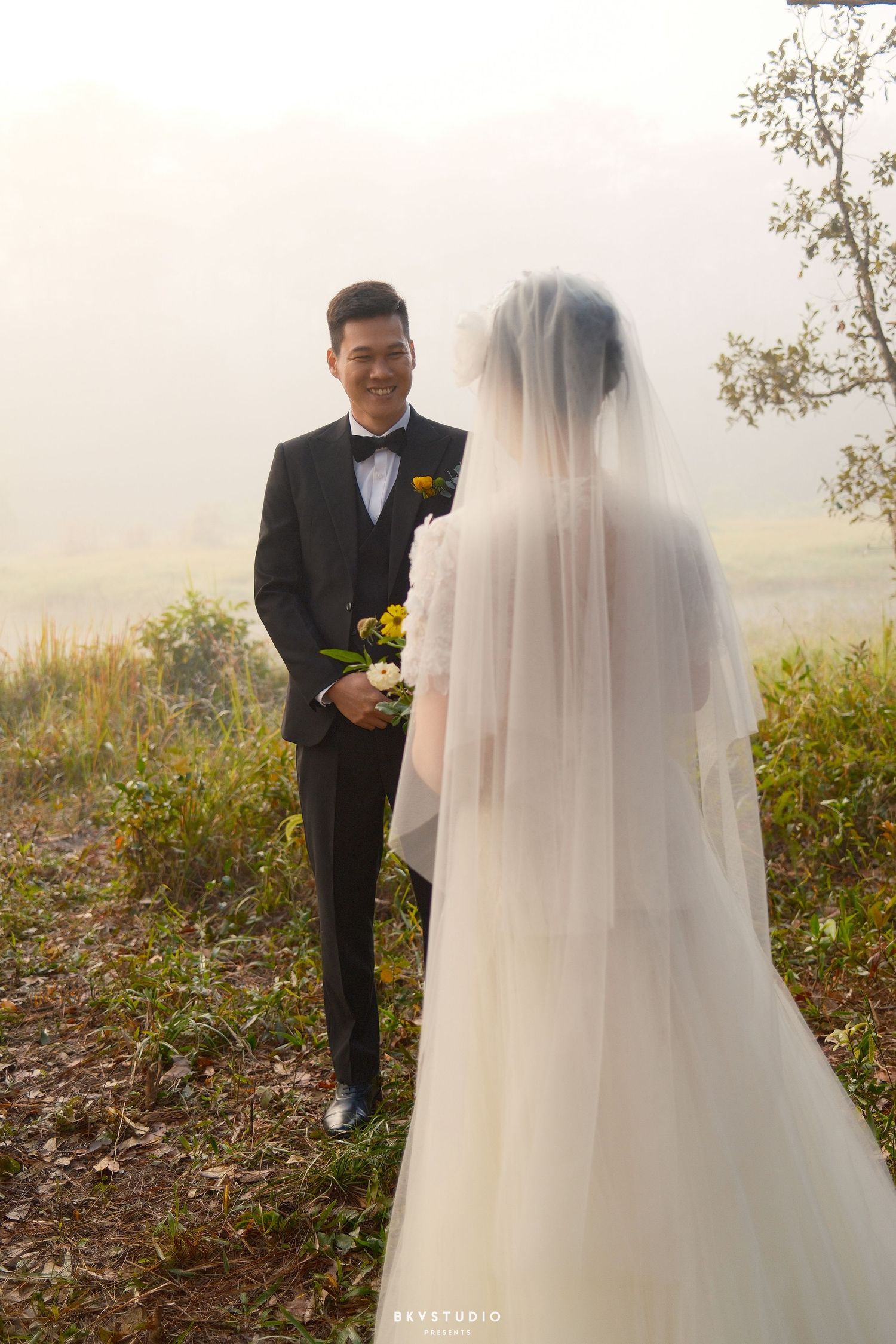 Groom in black tuxedo smiling at bride in white wedding dress and veil during outdoor ceremony.