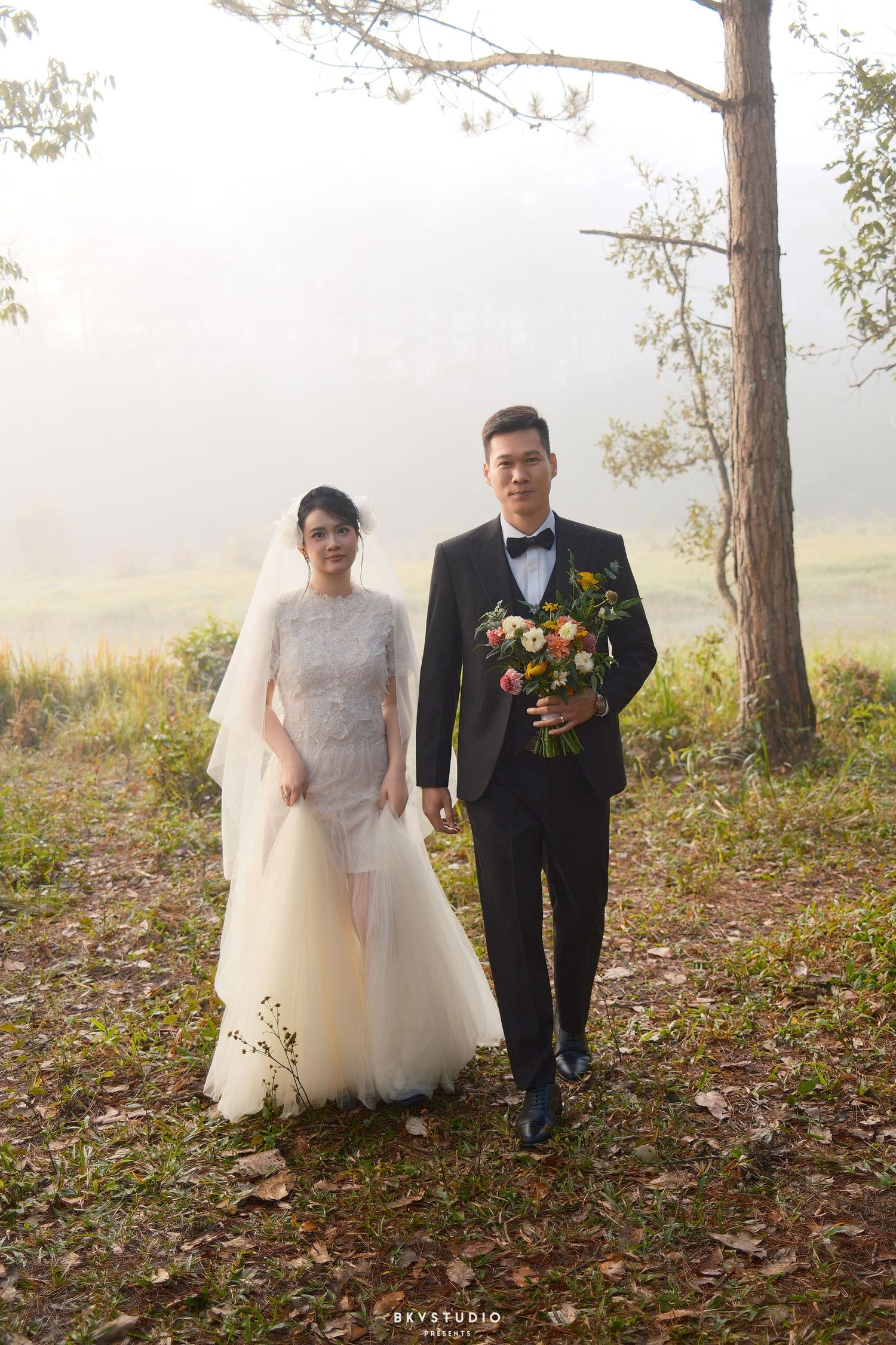 Bride and groom walk hand in hand through misty forest in elegant wedding attire with colorful bouquet.