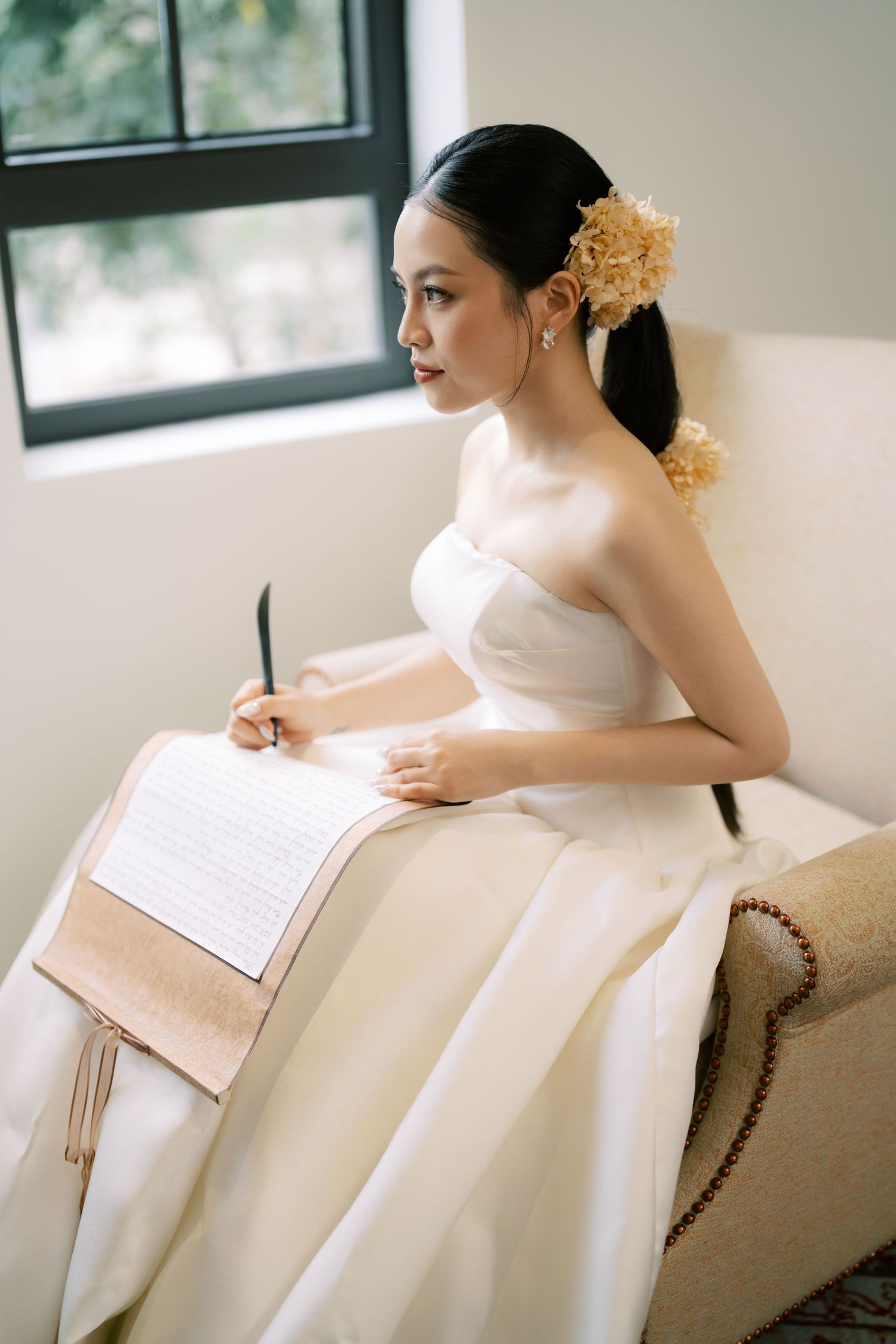 Bride in white strapless gown with gold hair flower writes vows on notepad while seated by window.