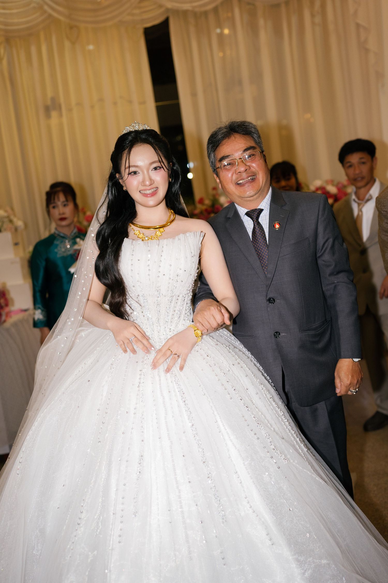 Bride in sparkling white ball gown with gold jewelry poses with older gentleman at wedding reception.