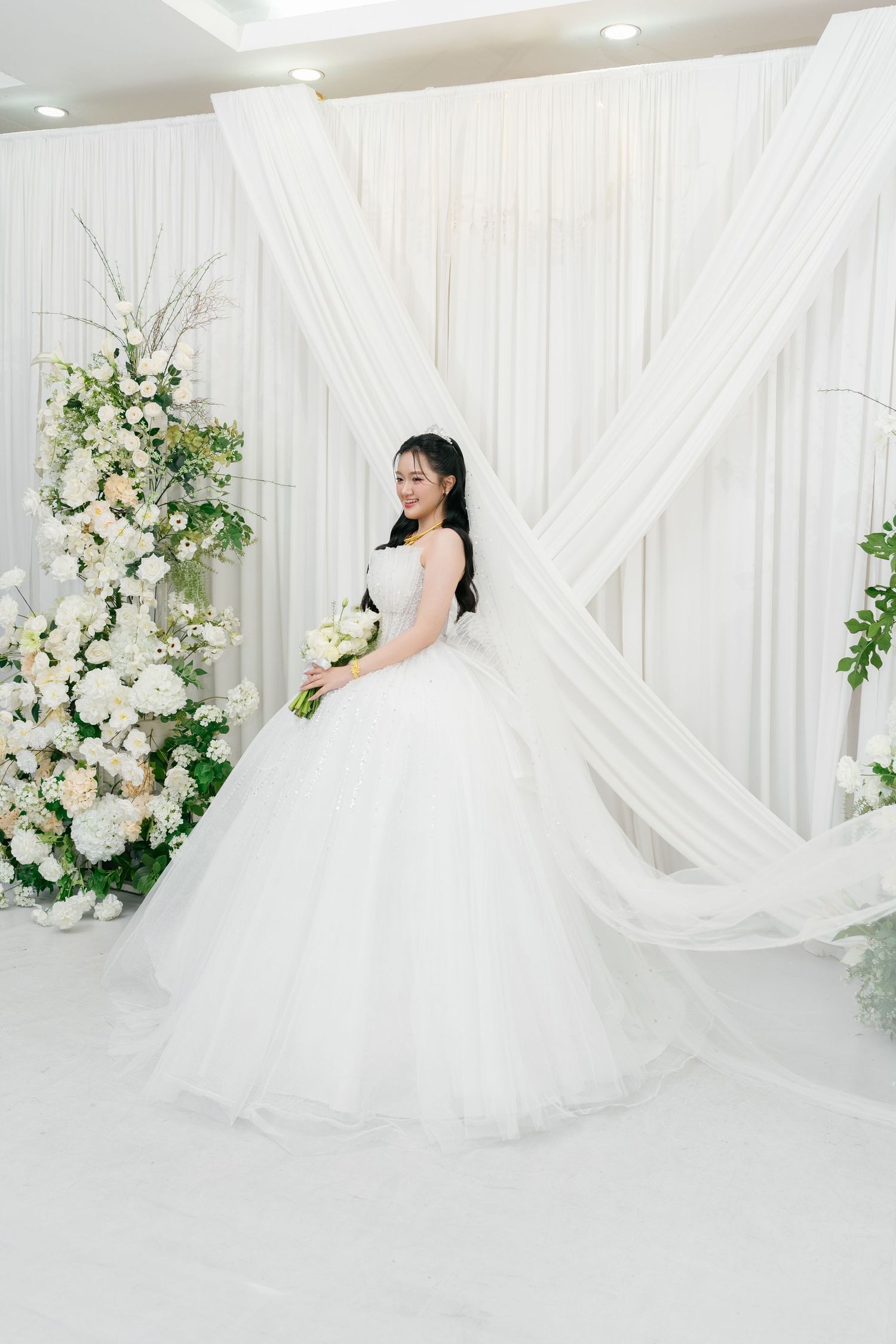 Elegant bride in a white ball gown holding a bouquet, posing before white drapes and floral arrangements.