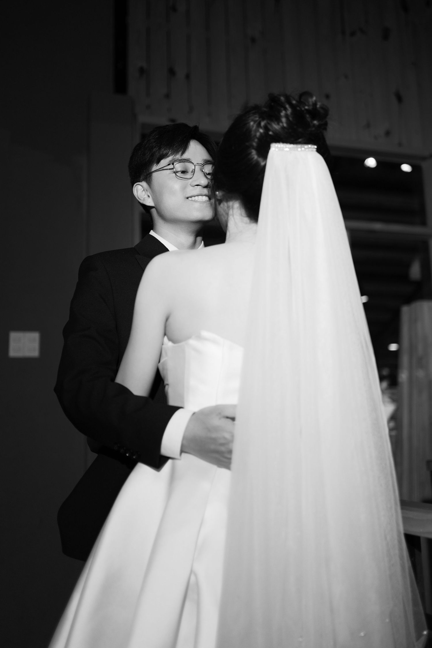 Black and white photo of a smiling groom embracing his bride in a wedding gown and veil.