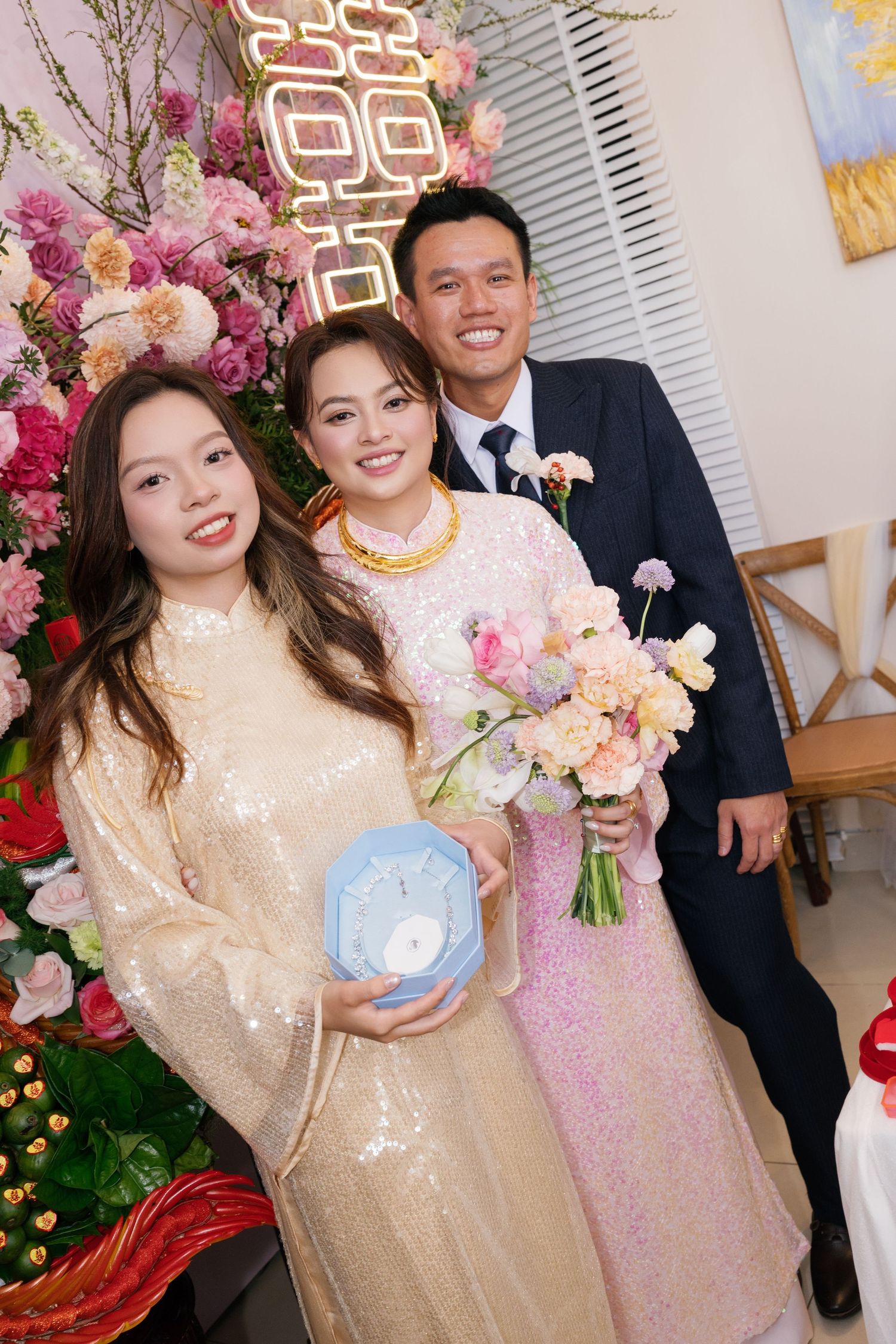Bride in pink sequin ao dai with gold jewelry holds bouquet posing with guests at Vietnamese wedding.