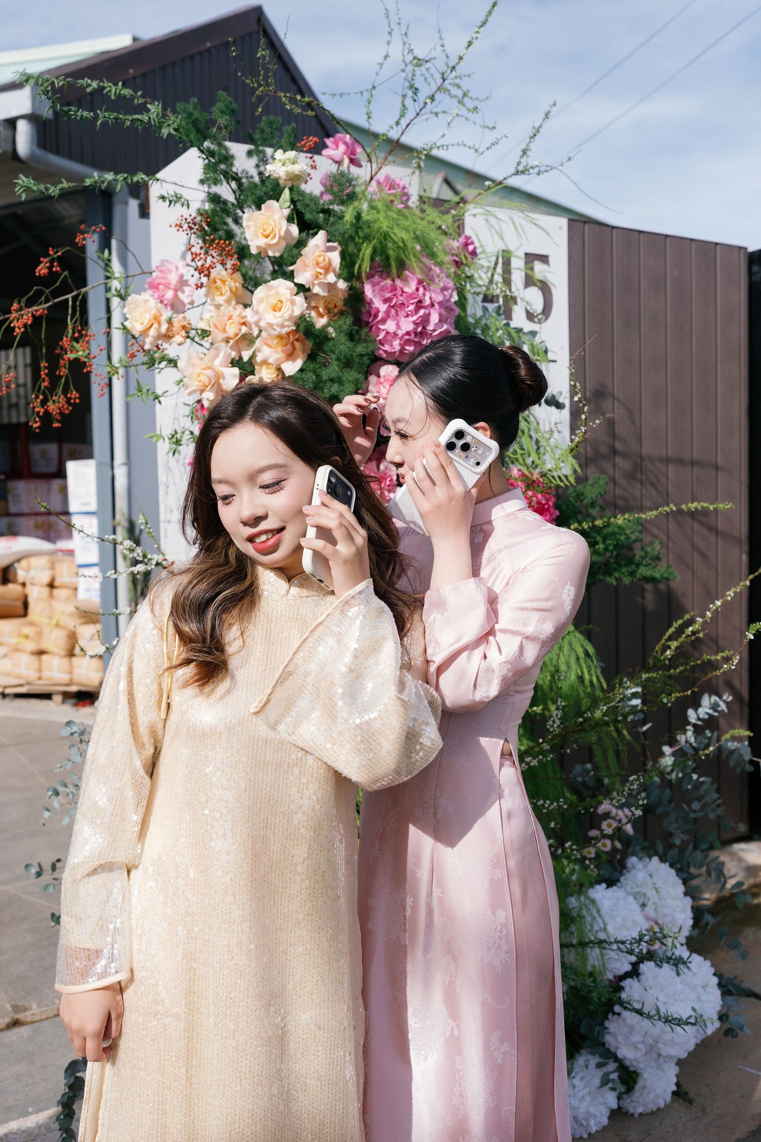 Two women in elegant ao dai dresses look at smartphones near a colorful floral arrangement outdoors.