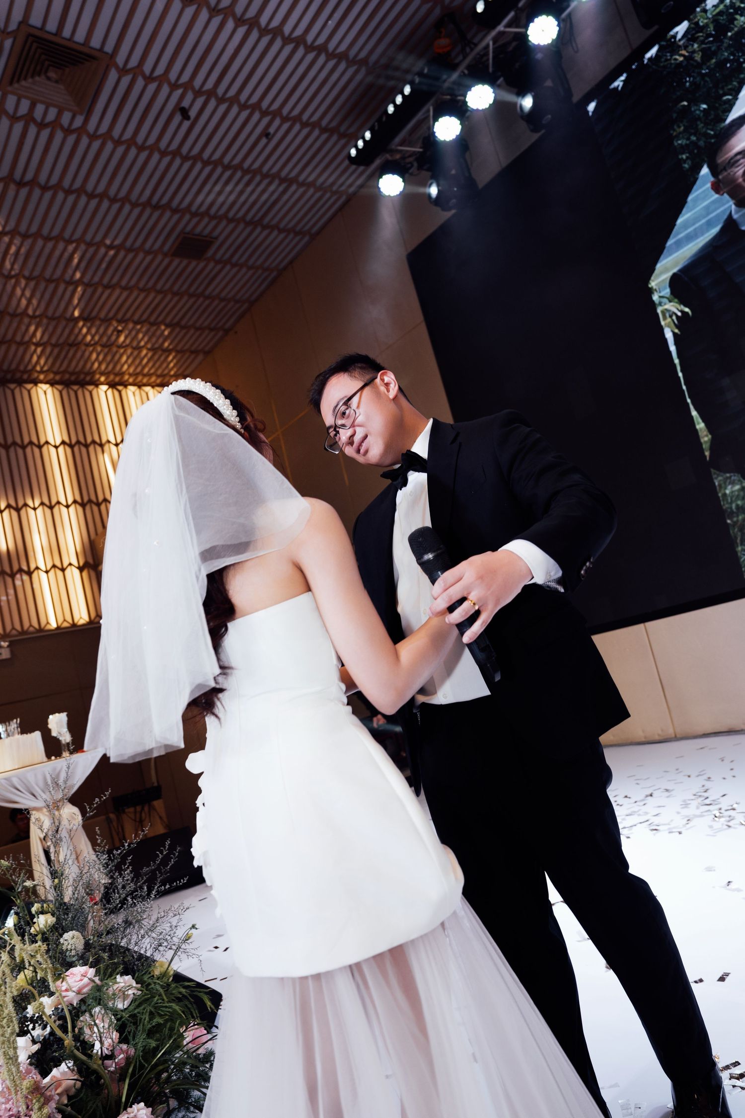 Groom in black tuxedo holds microphone while embracing bride in white dress during wedding reception dance.
