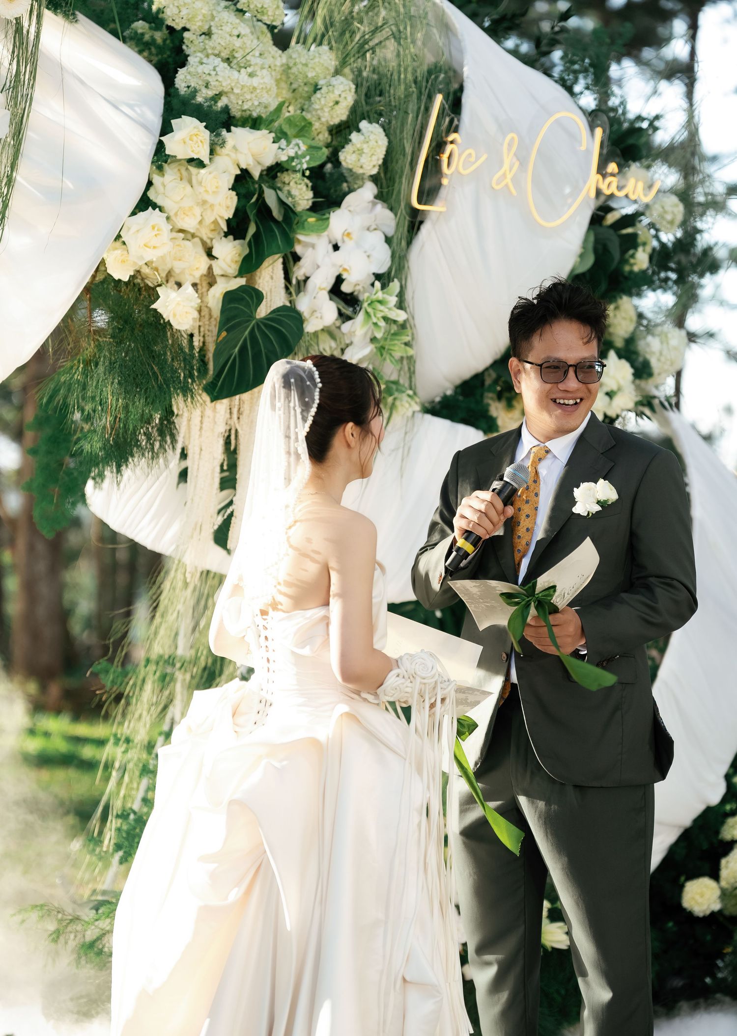 A groom in a gray suit reads vows to his bride in a white wedding dress at an outdoor ceremony.