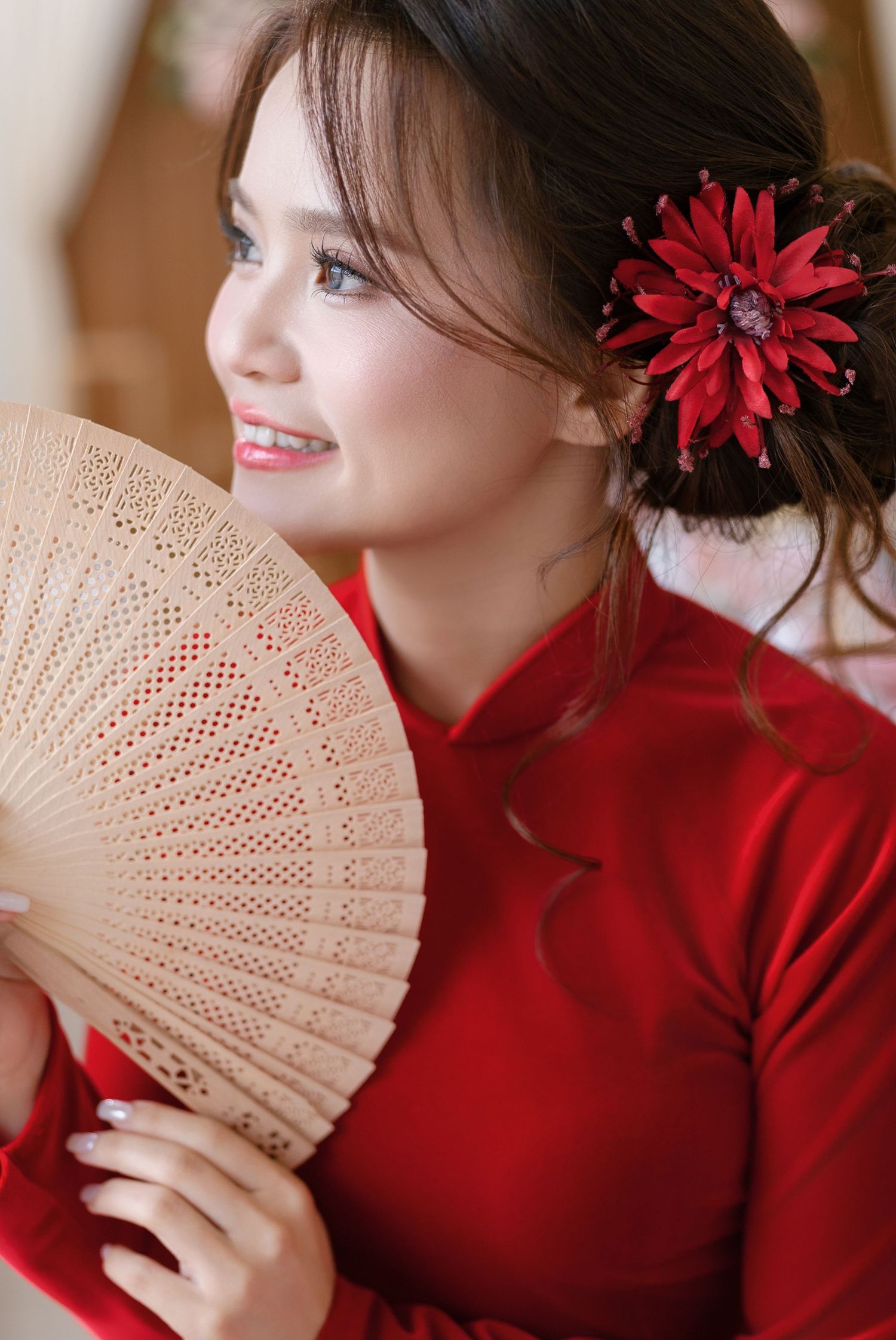Young woman in red ao dai holding a decorative wooden fan with a red flower hair accessory, smiling gracefully.