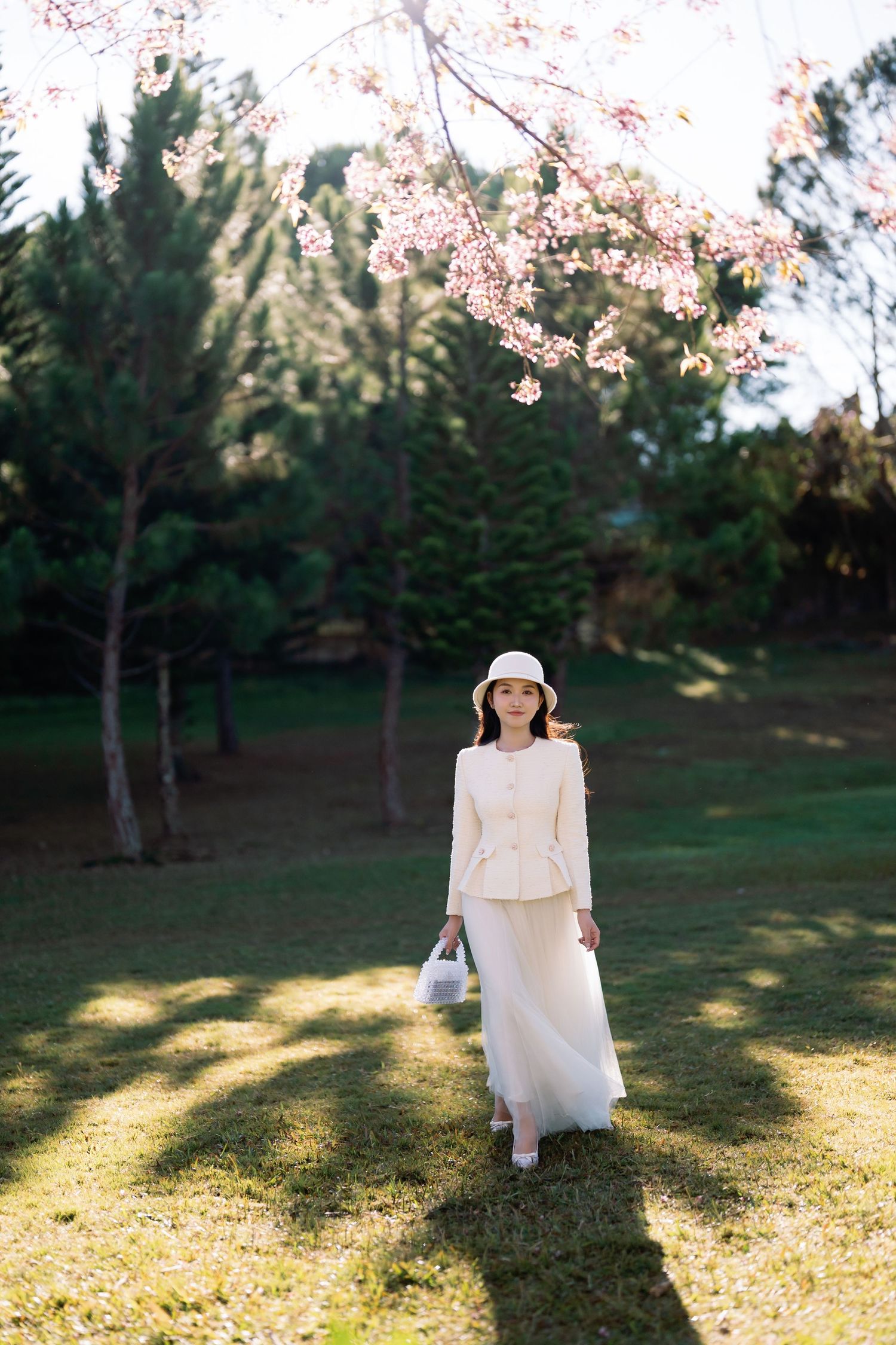 Woman in white outfit and hat stands under cherry blossom tree in sunny green park.