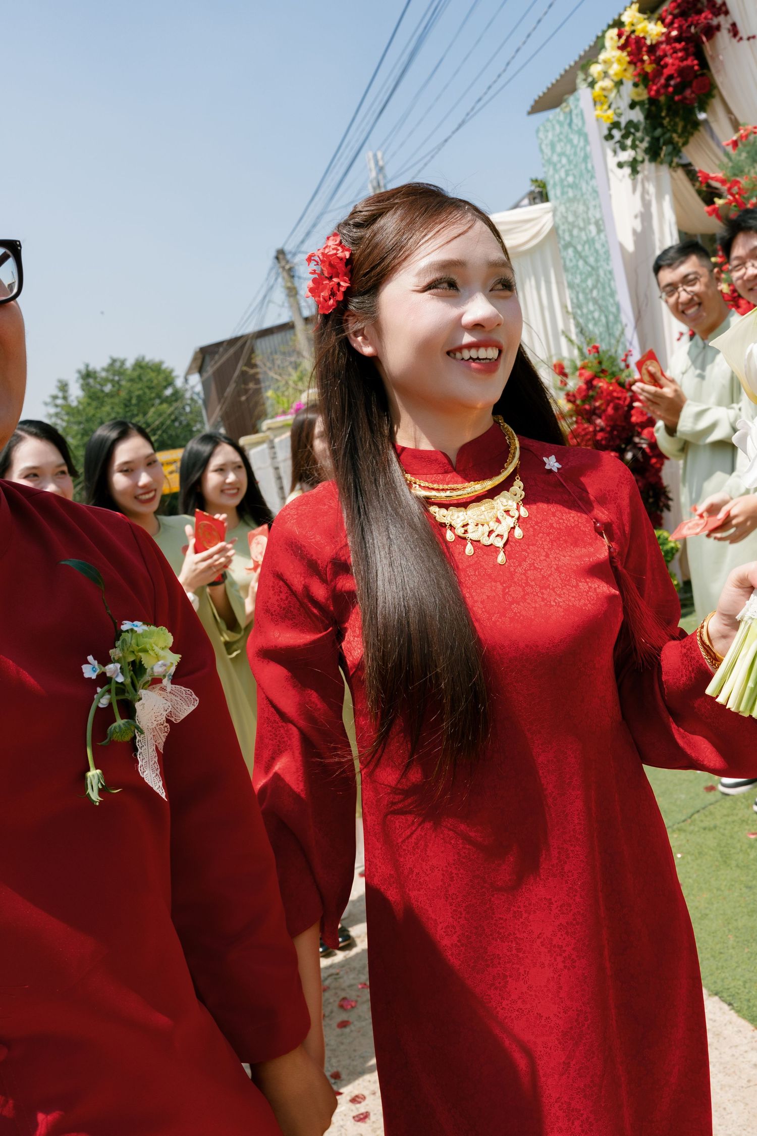 Smiling young woman in red traditional Vietnamese ao dai dress with gold jewelry at an outdoor wedding ceremony.