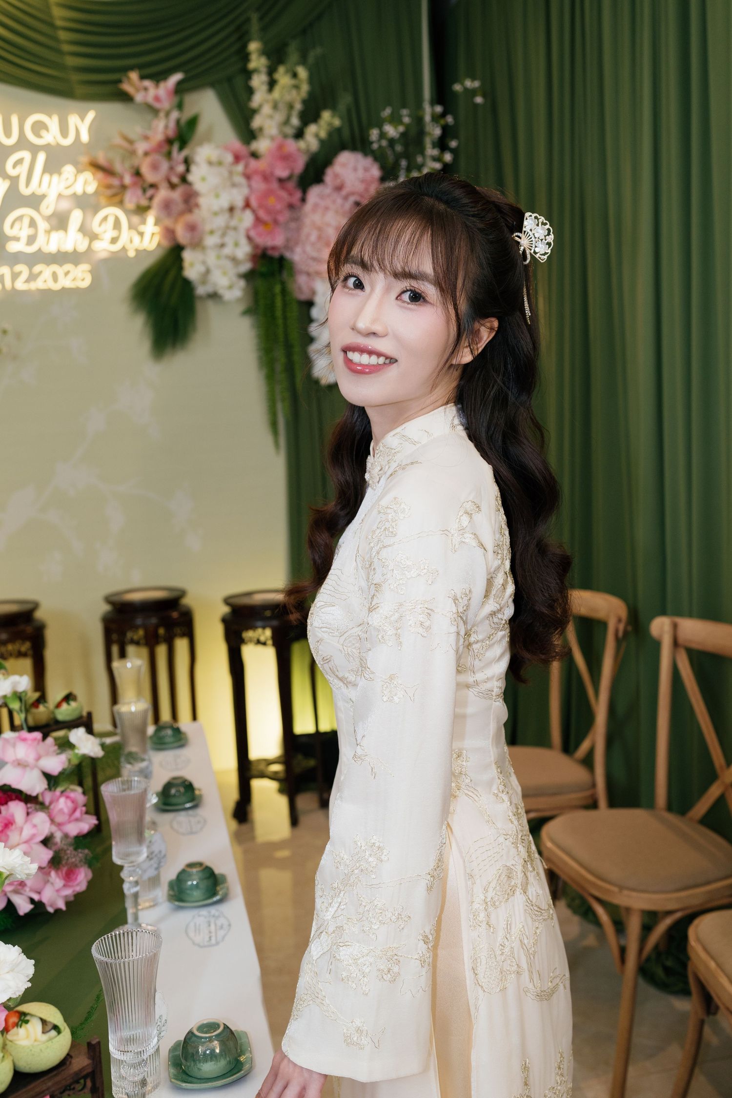 A smiling bride in a white floral ao dai dress poses at a Vietnamese wedding ceremony with pink flowers and green decor.