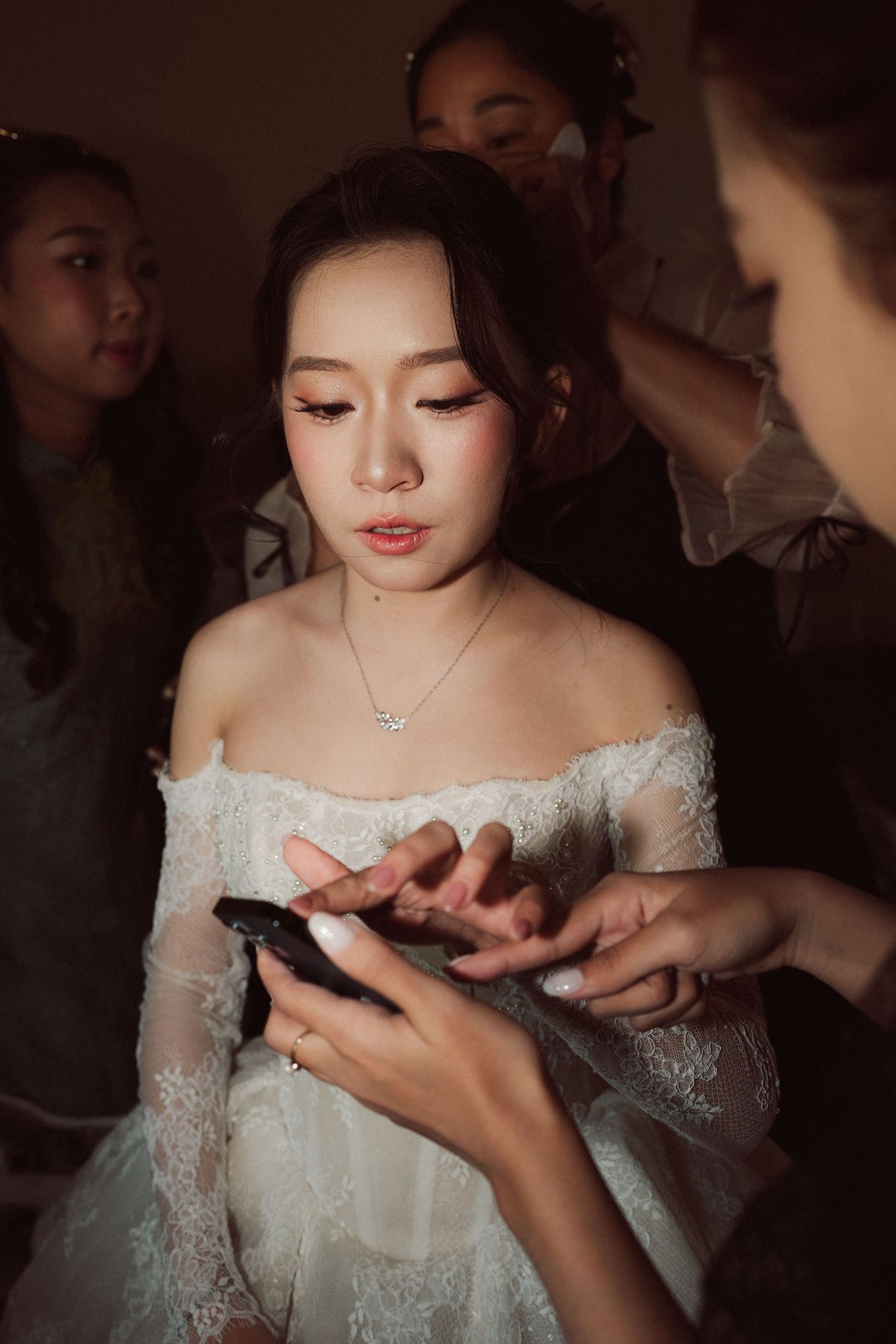Bride in white off-shoulder lace dress with star necklace looks at phone while attendants help prepare for wedding.