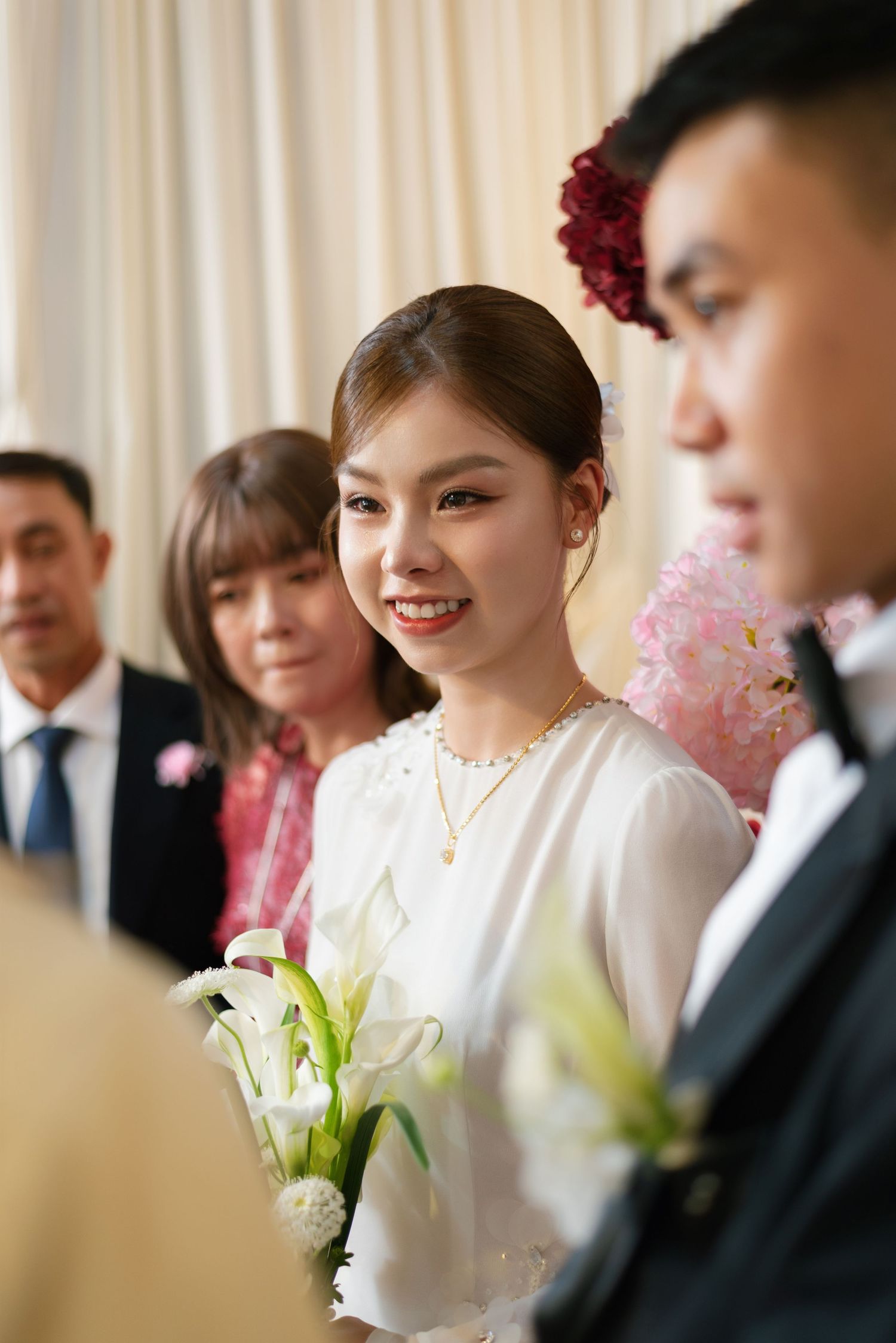 Smiling Asian bride in white dress holding white calla lilies with red flower hair accessory at wedding ceremony.