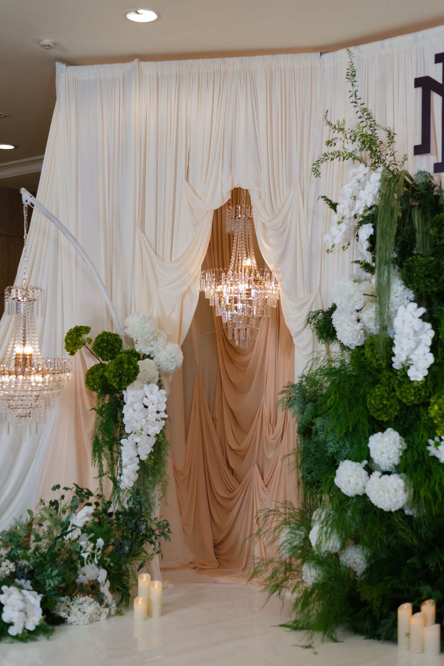 Elegant wedding backdrop with white draping, crystal chandeliers, and lush green and white floral arrangements.