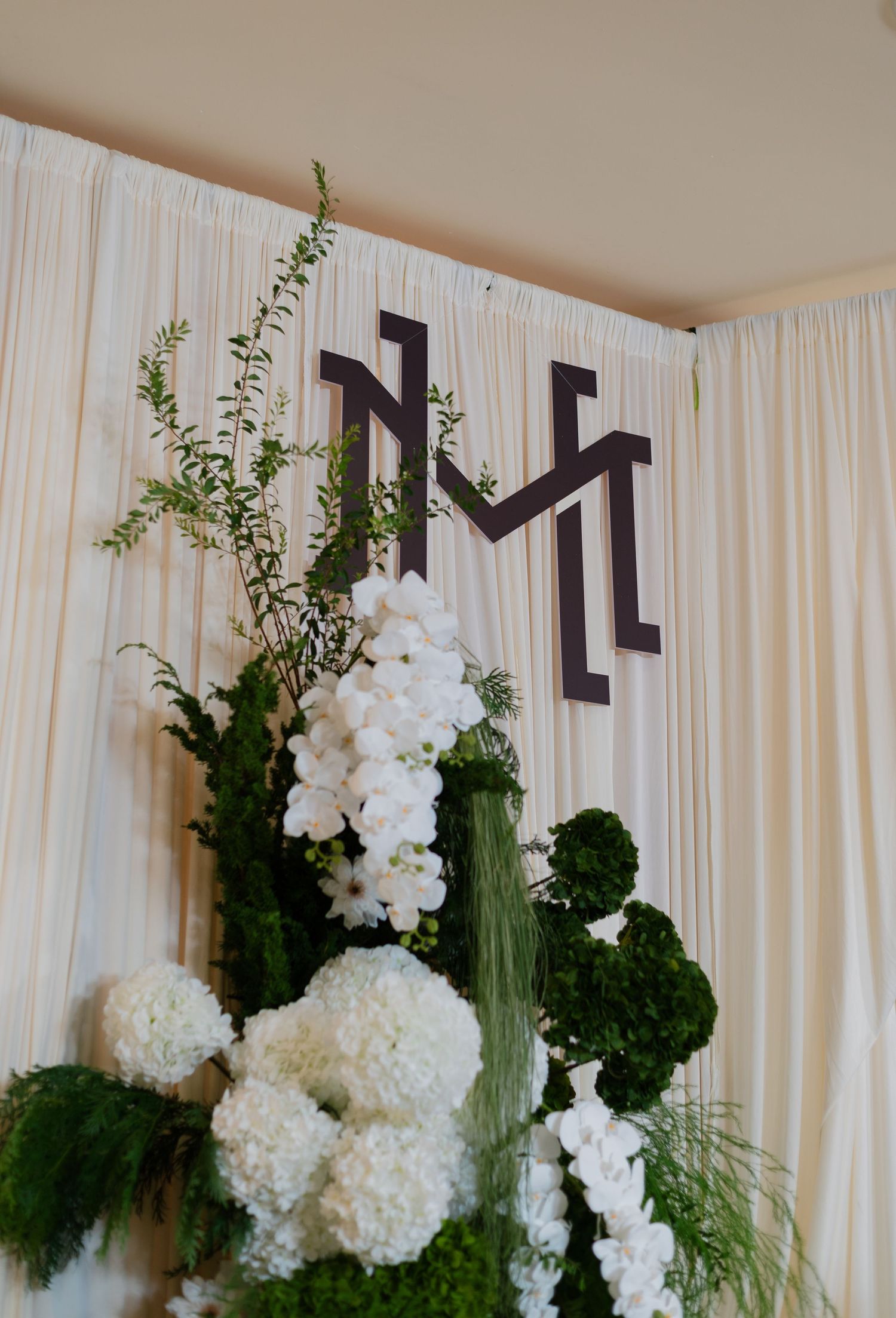 Elegant wedding backdrop with white draping, monogram letters, and lush white floral arrangements with green foliage.