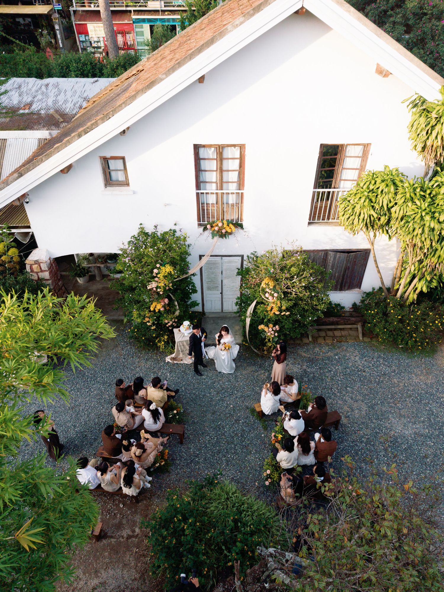 Aerial view of an intimate outdoor wedding ceremony in a courtyard beside a white chapel surrounded by lush greenery.