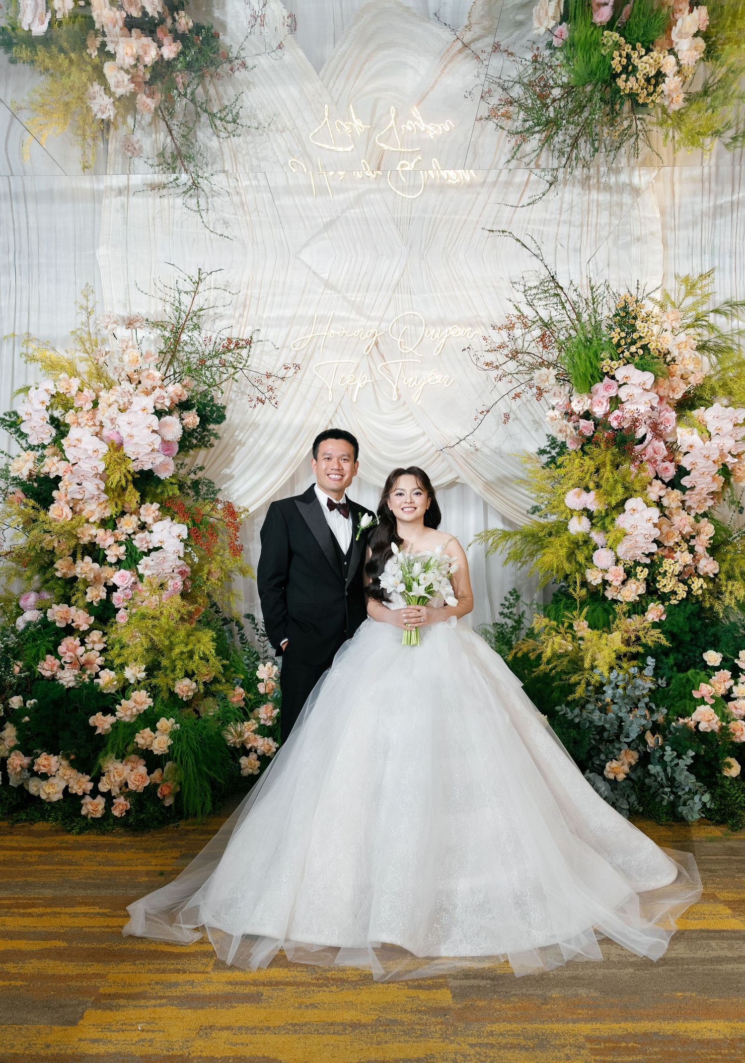 Bride and groom posing before a floral arch with pink blooms and greenery at an elegant wedding reception.