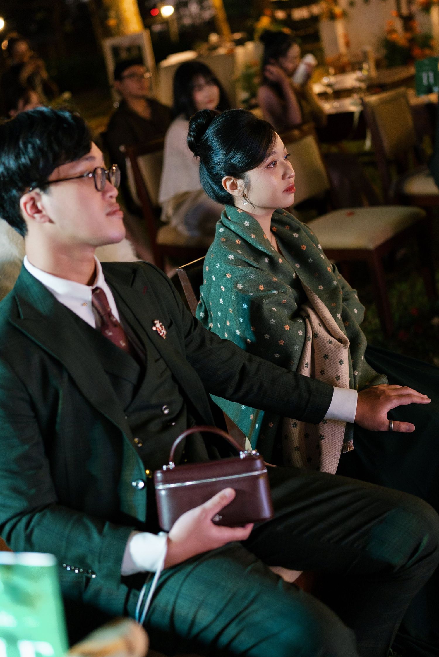 Elegantly dressed wedding guests in dark green suits watching ceremony at outdoor evening reception.