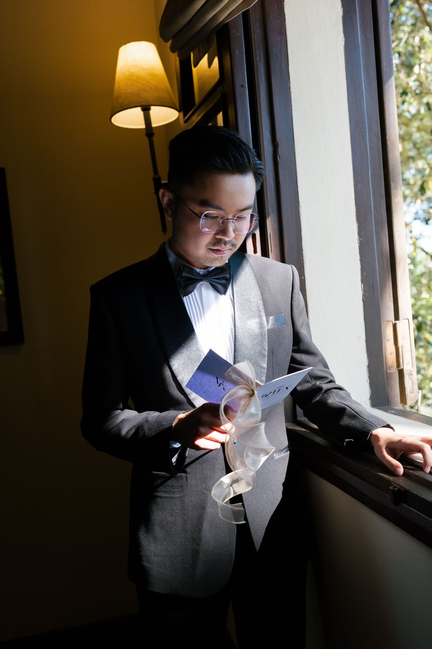Groomsman in gray suit reading a wedding card while standing by a window indoors.